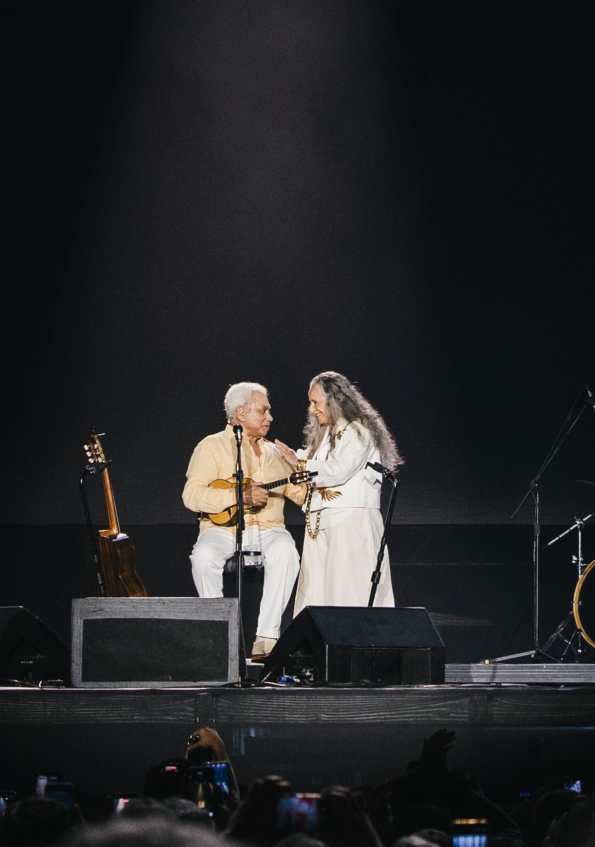 Two musicians share an intimate moment on stage, one playing guitar, both dressed in white under a spotlight.