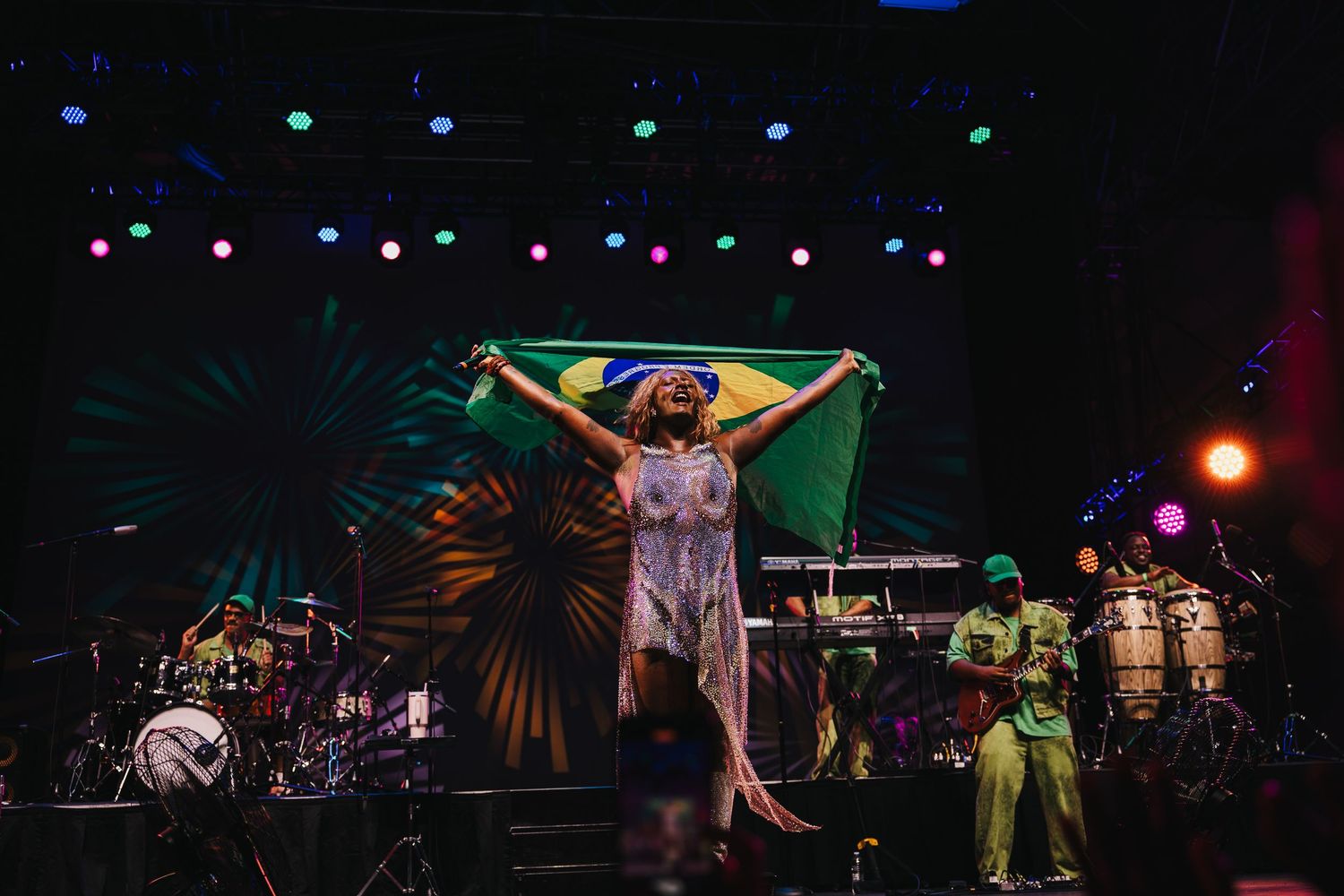 A performer holds a Brazilian flag triumphantly on a colorful stage during a live music performance.