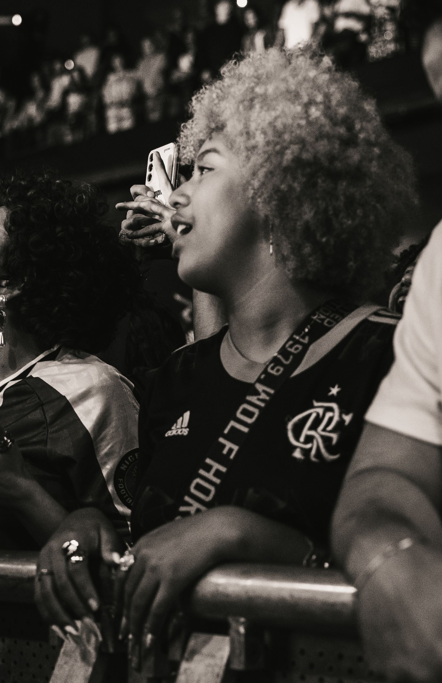 Black and white photo of a curly-haired fan singing along at a concert holding a phone.