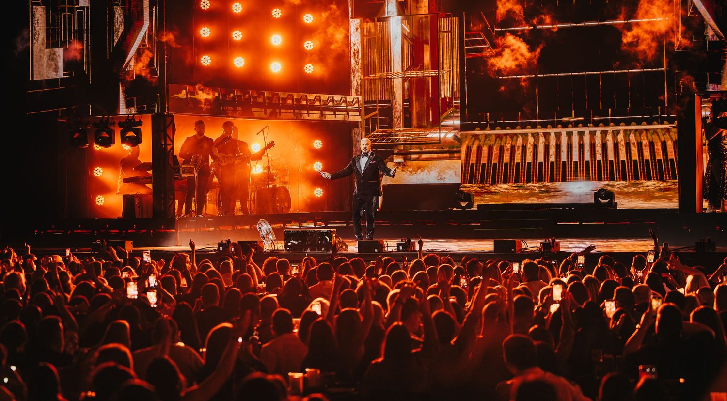 Live concert stage with bright orange lights, performers, and an energetic crowd raising hands in a packed venue.