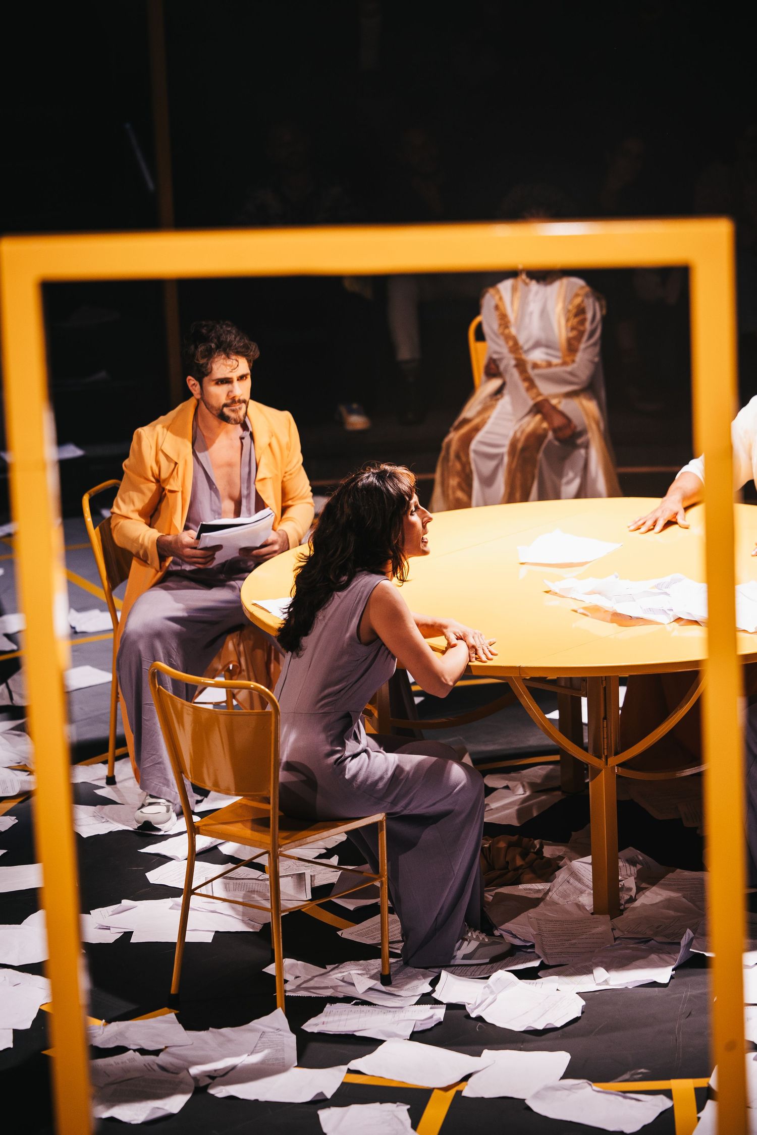 Two actors sit at a yellow table on a theatrical stage scattered with papers, in a dramatic scene.