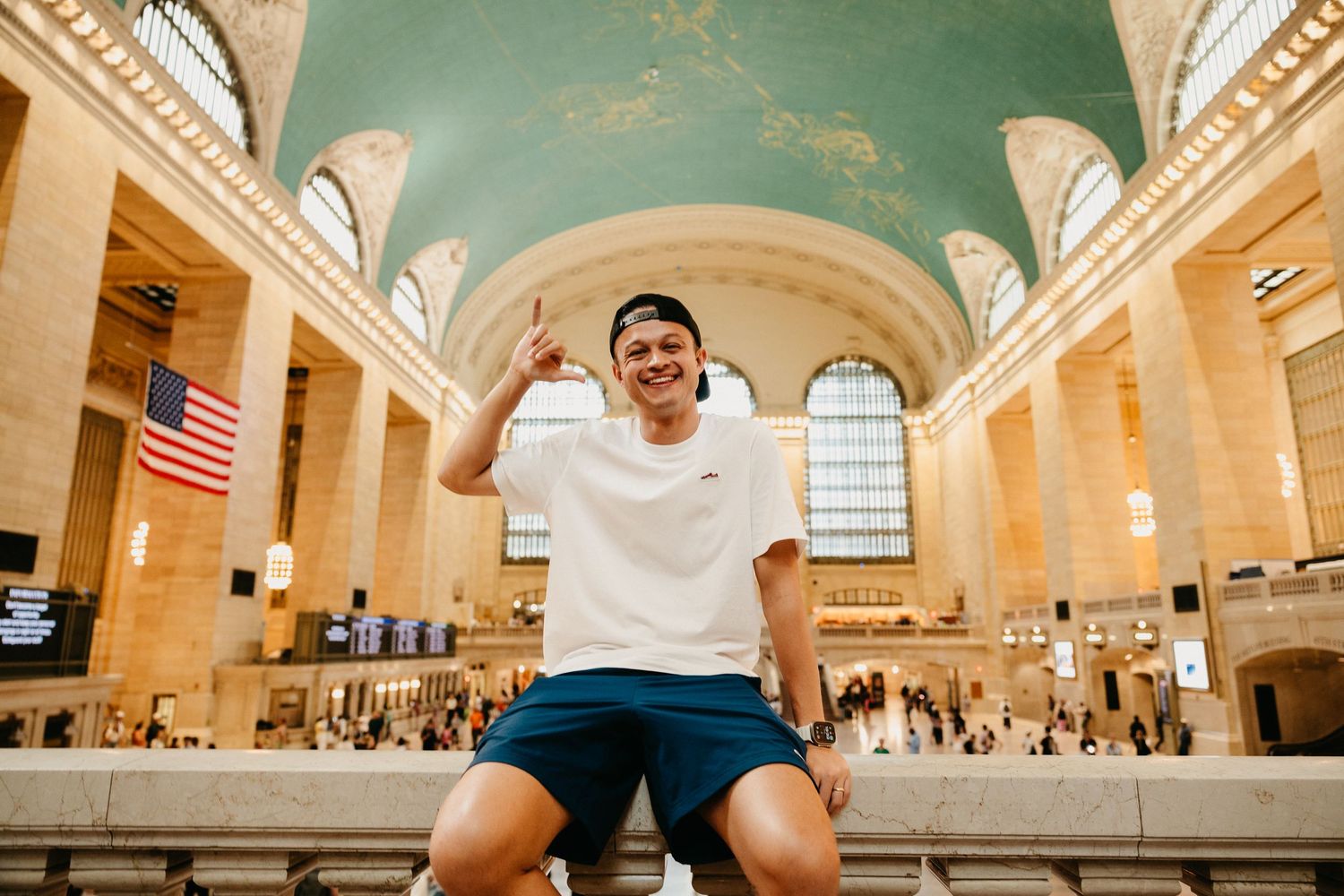 Smiling man in white tee and blue shorts posing at Grand Central Terminal NYC.
