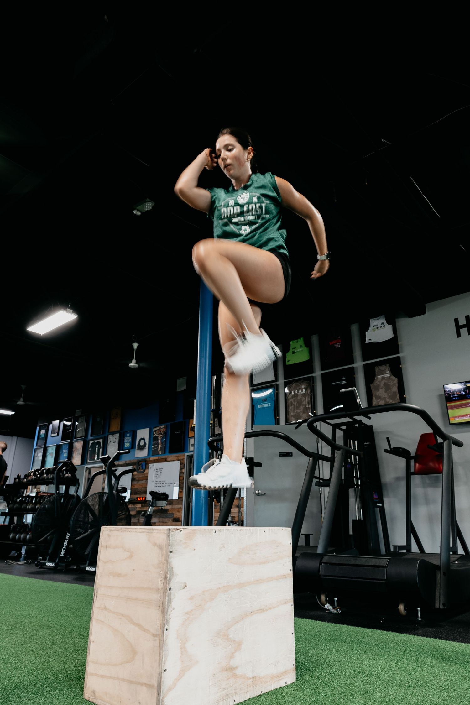 Woman performing a box jump exercise on a wooden plyo box in a CrossFit gym wearing a green tank top.