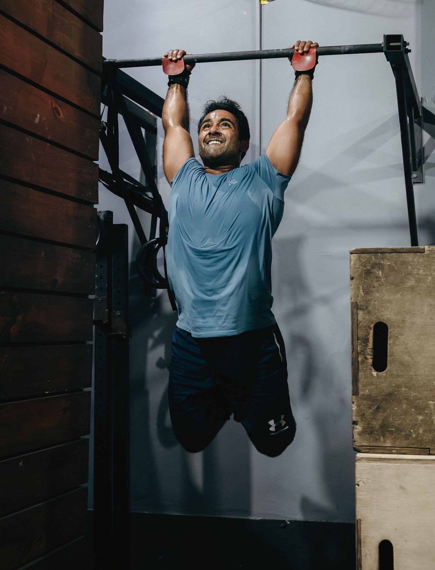 Smiling man in blue shirt and black shorts doing pull-ups on a bar in a gym.