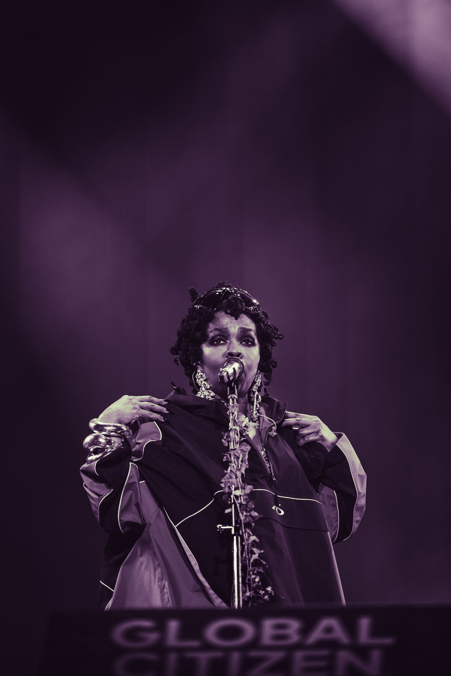 A performer sings on stage at a Global Citizen event under purple stage lighting, wearing a floral crown and dark outfit.