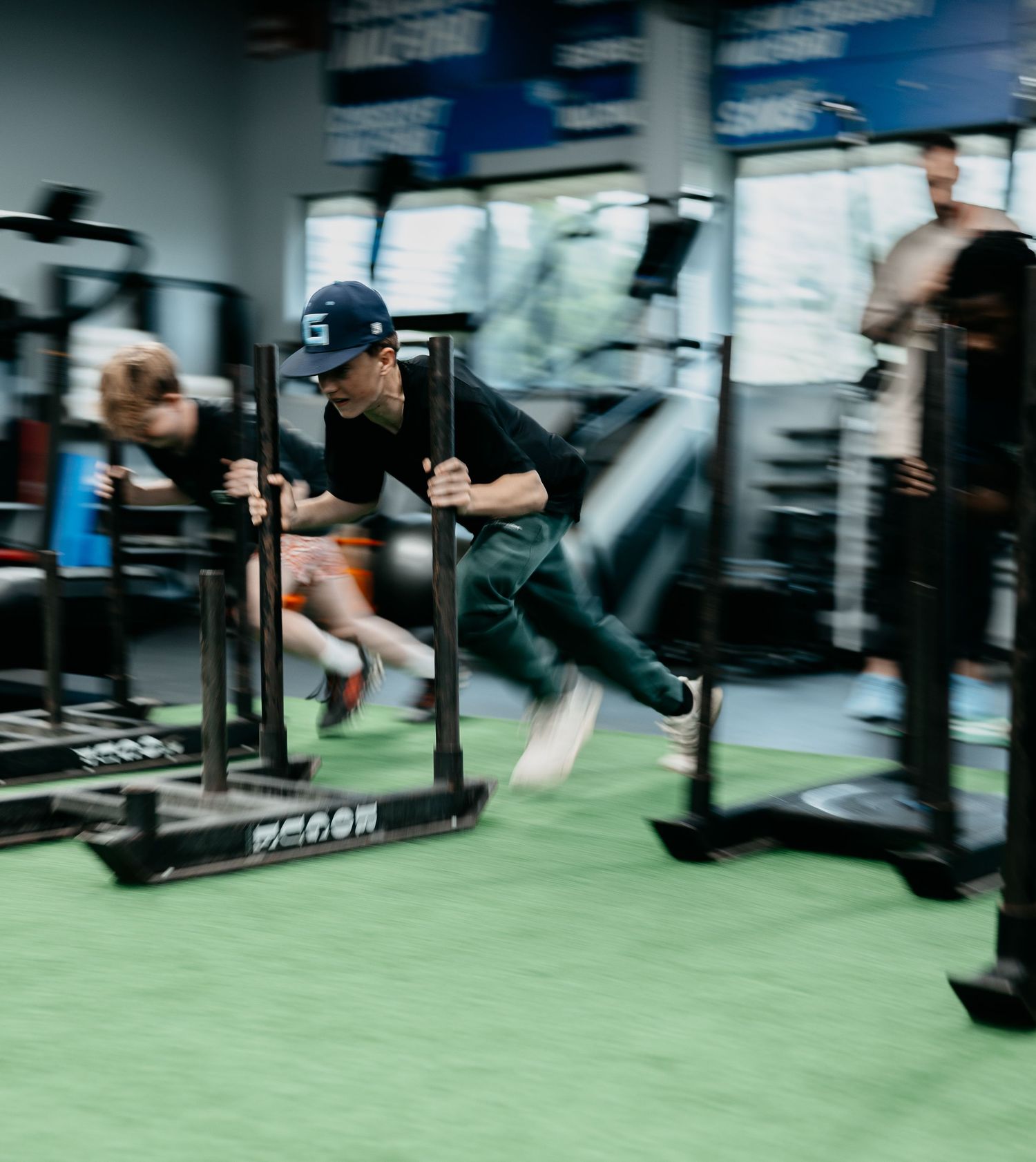 Athletes push weighted sleds across green turf in a modern gym during an intense speed and strength training session.