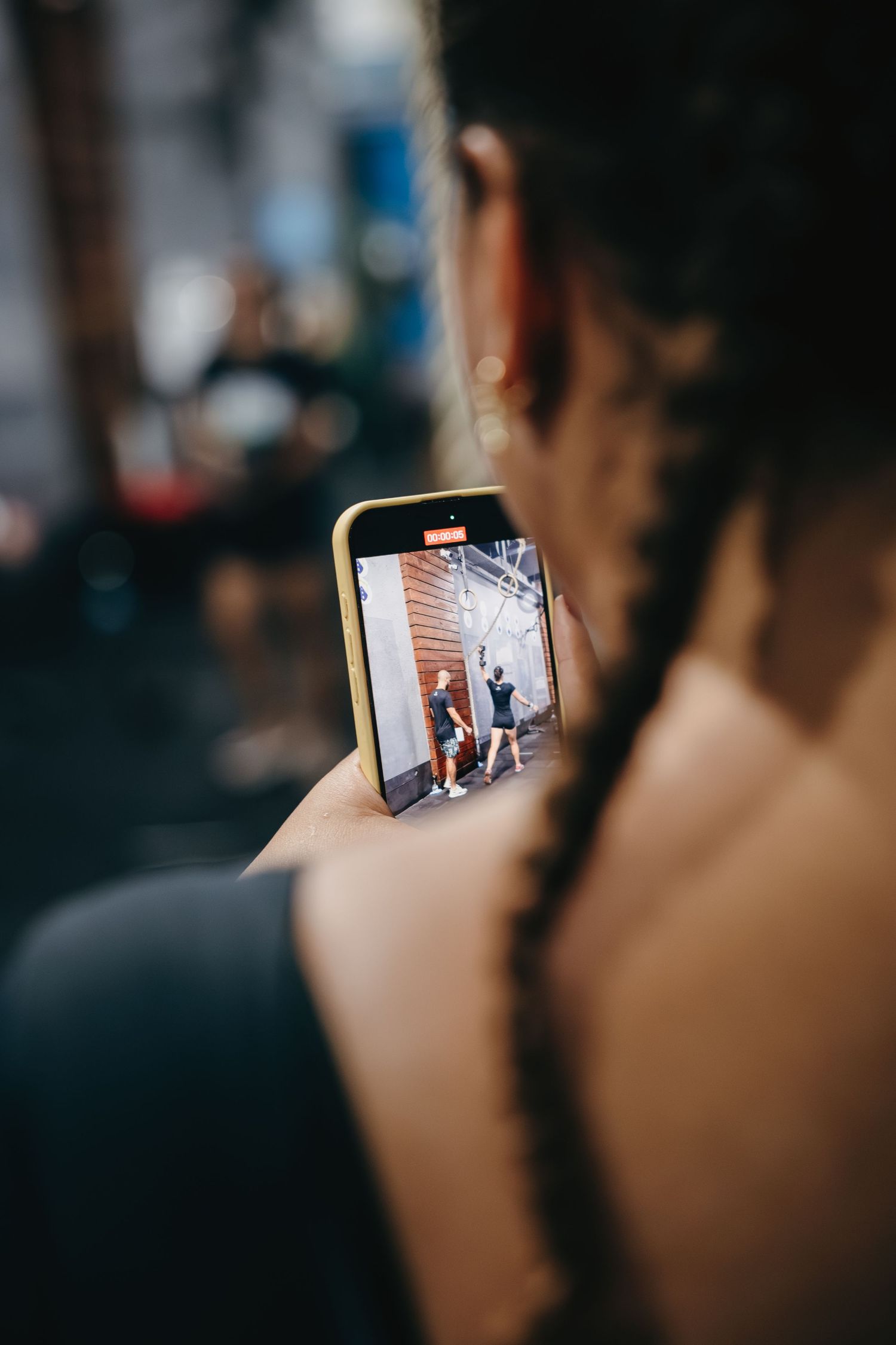 Woman with braids filming people on her yellow-cased smartphone in a gym setting.