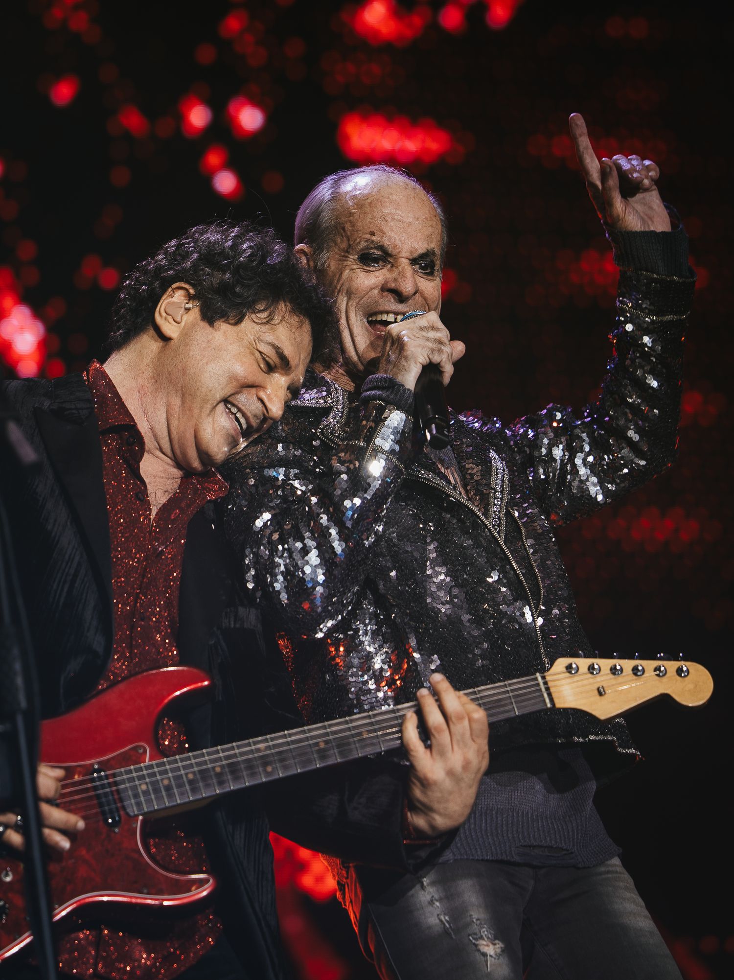 Two male rock musicians perform together on stage, one playing guitar under red concert lighting.