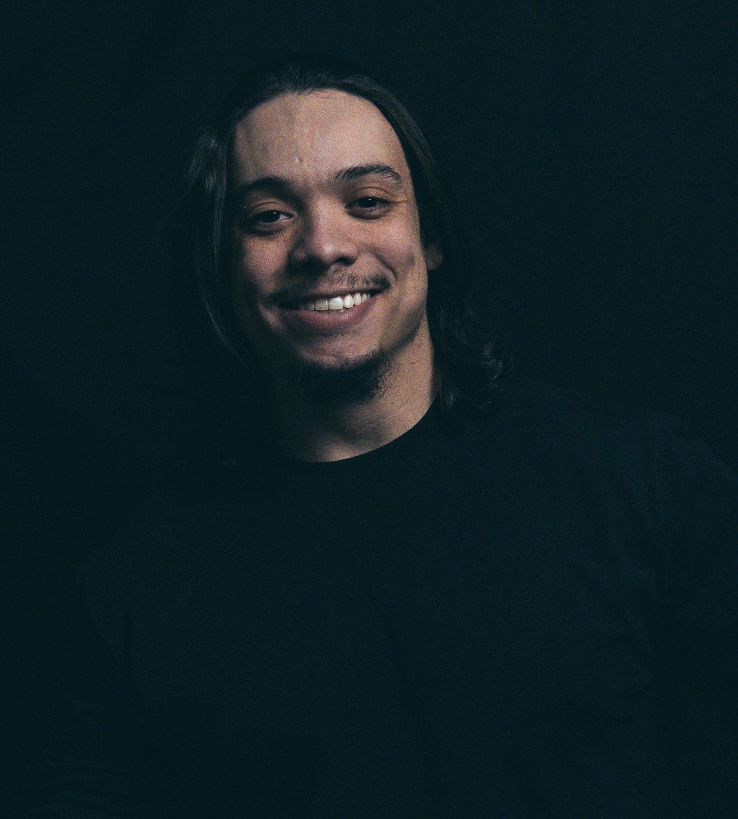 Young man with long hair smiling against a dark background in a black shirt.