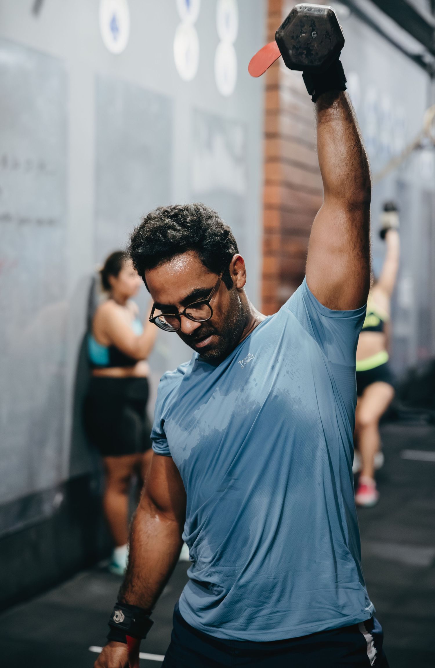 A man in a blue shirt lifts a dumbbell overhead with one arm during a gym workout session.