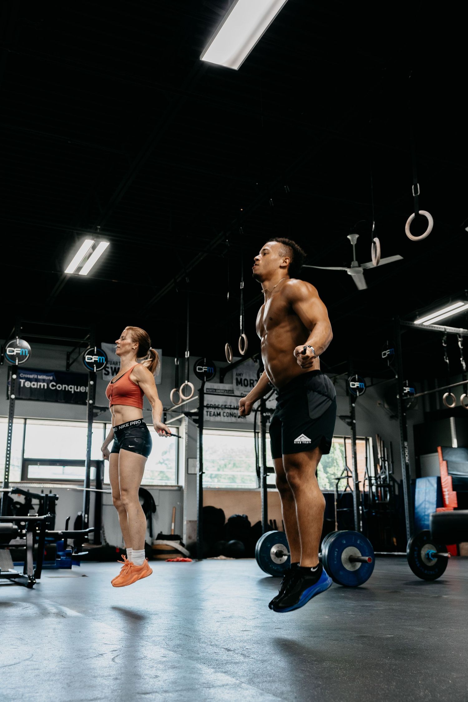 Two athletes jump rope together in a CrossFit gym, with barbells and gymnastic rings visible in the background.