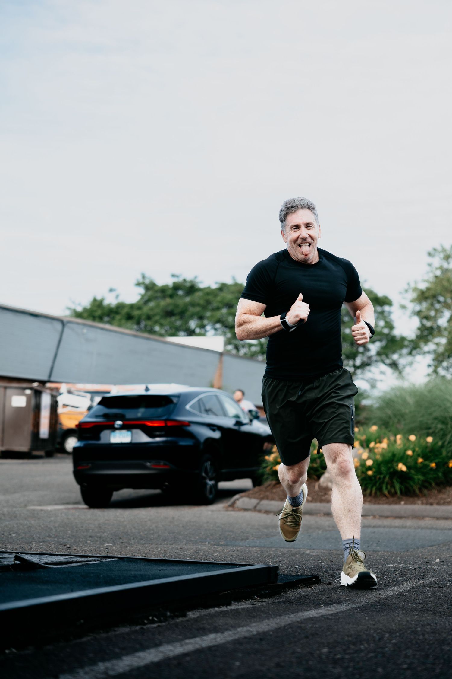 Middle-aged man in black shirt and shorts running outdoors in a parking lot, smiling during a daytime workout.
