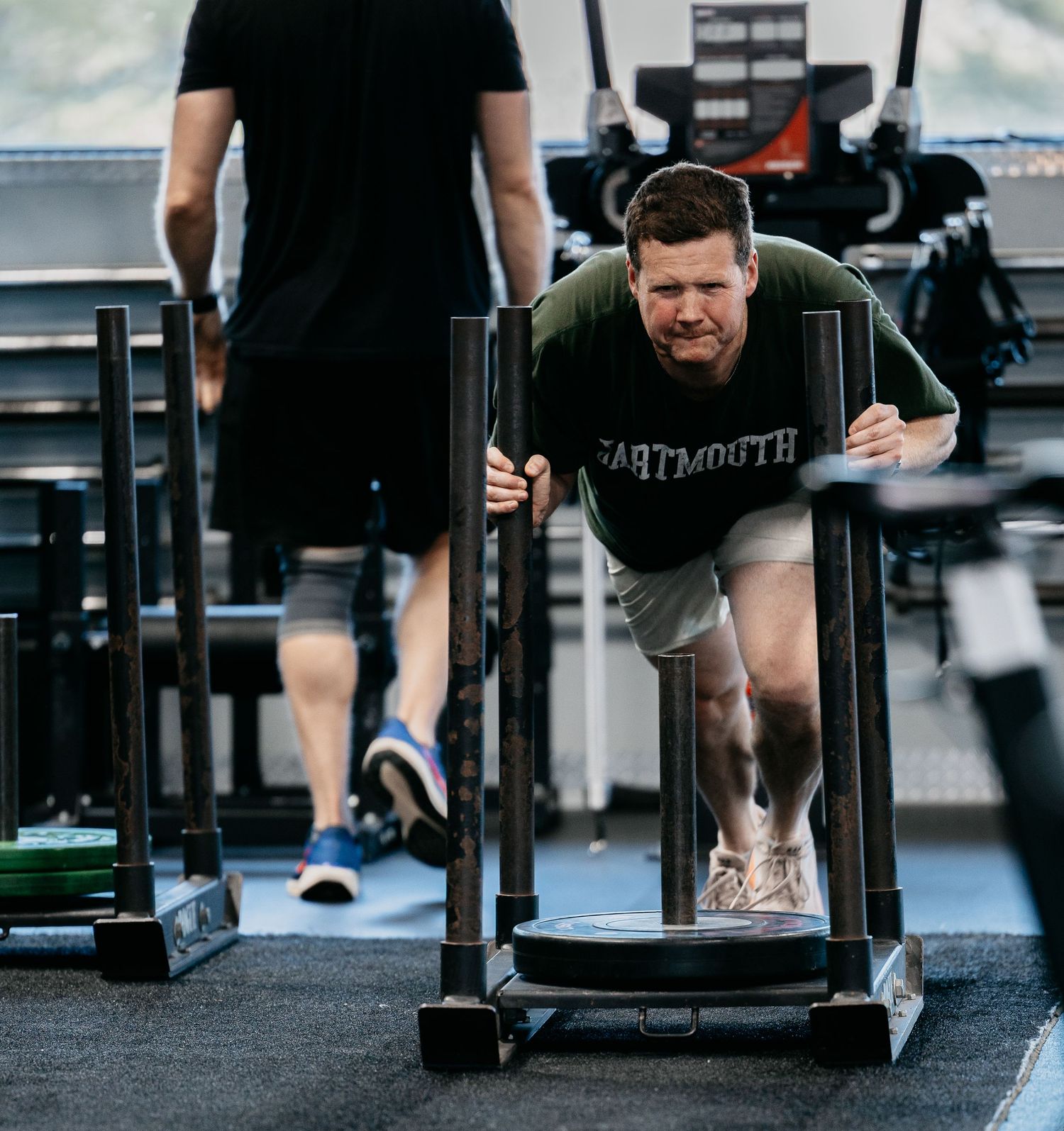 Man in Dartmouth shirt pushing a weighted sled at a CrossFit gym during an intense workout.