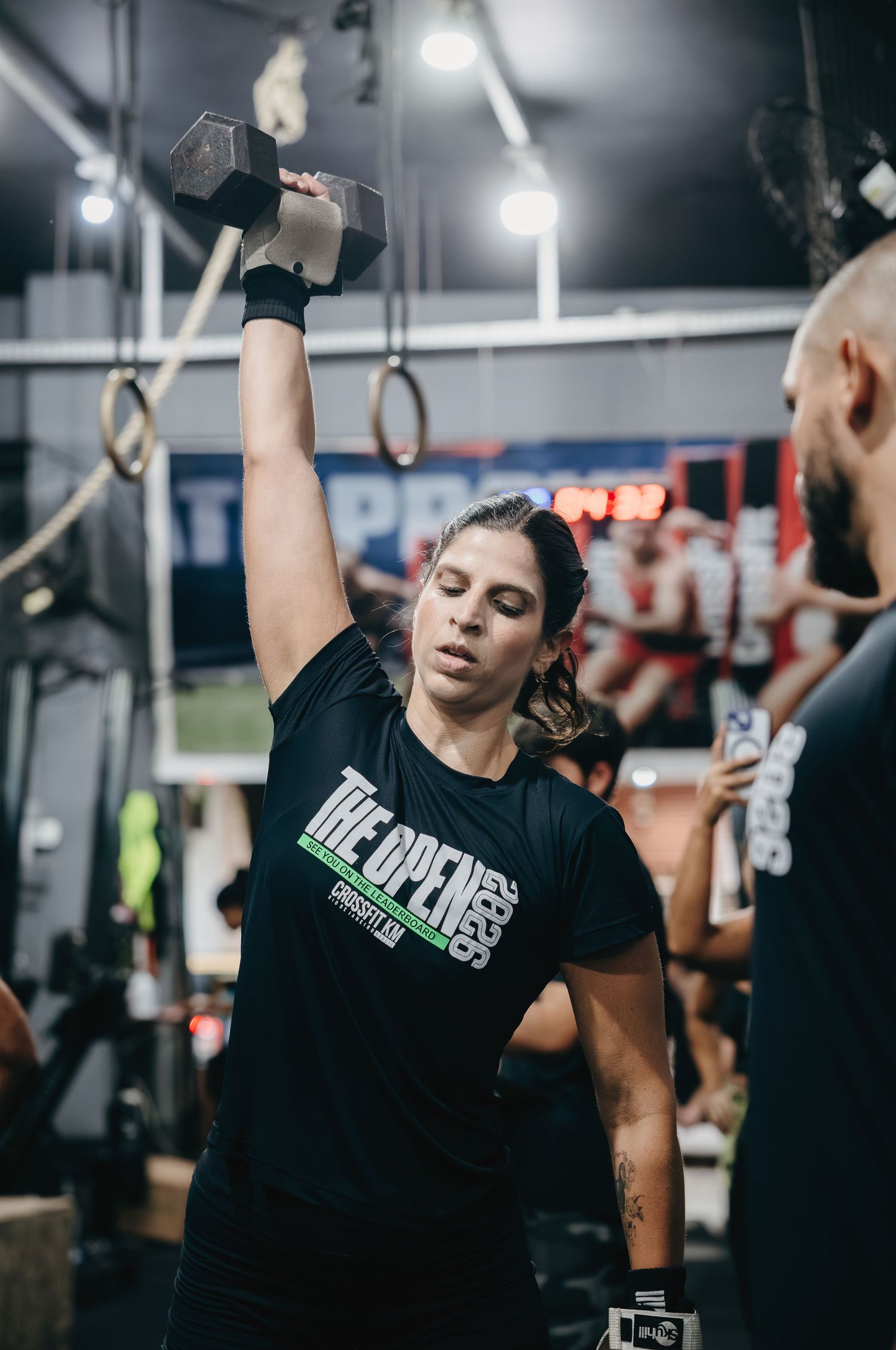 Woman in CrossFit Open shirt performs a single-arm dumbbell overhead press during a gym competition.