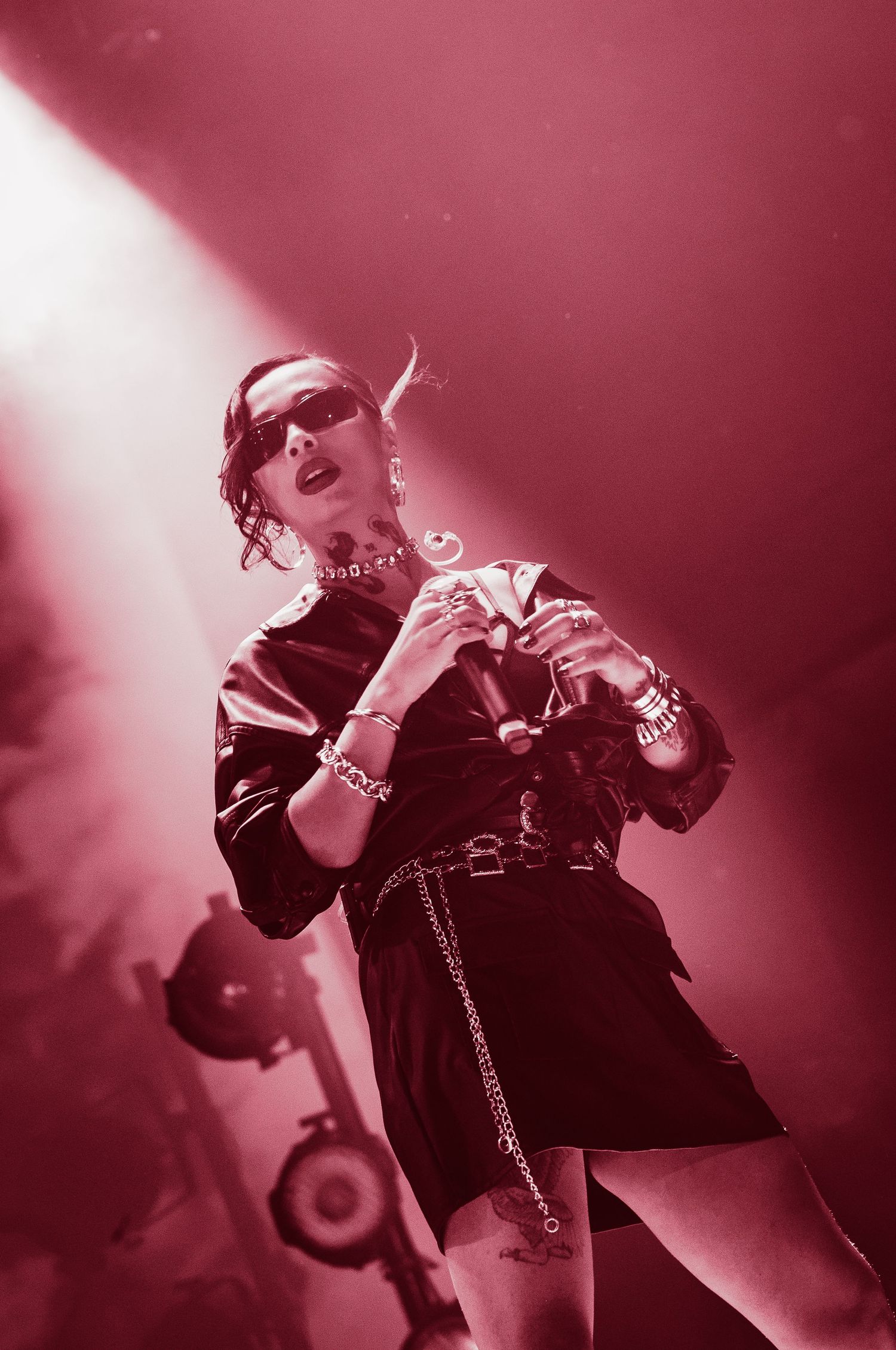 Female performer singing on stage under red dramatic lighting wearing leather outfit and sunglasses.