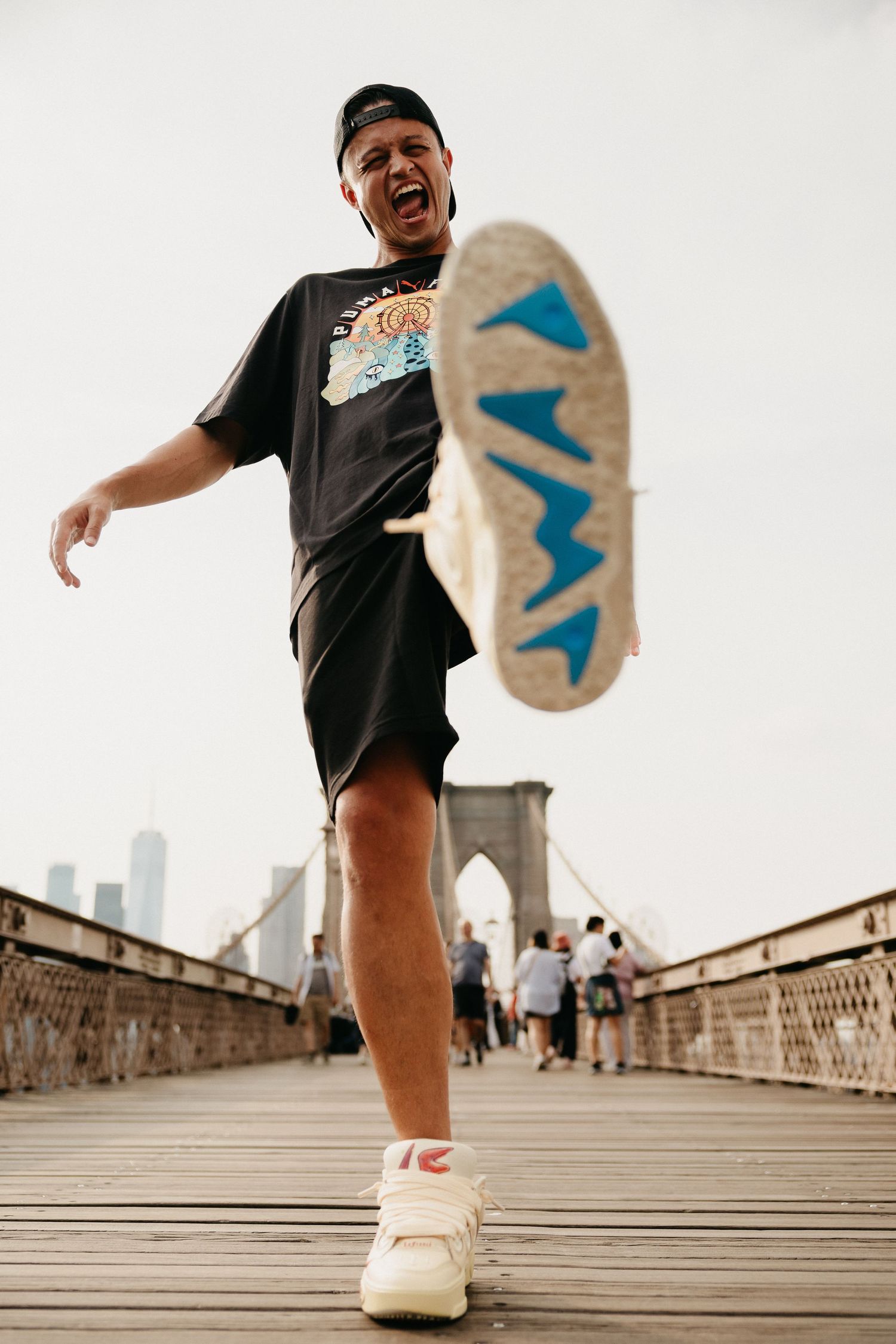 Man kicking his shoe toward camera on Brooklyn Bridge wearing black shorts and graphic tee.
