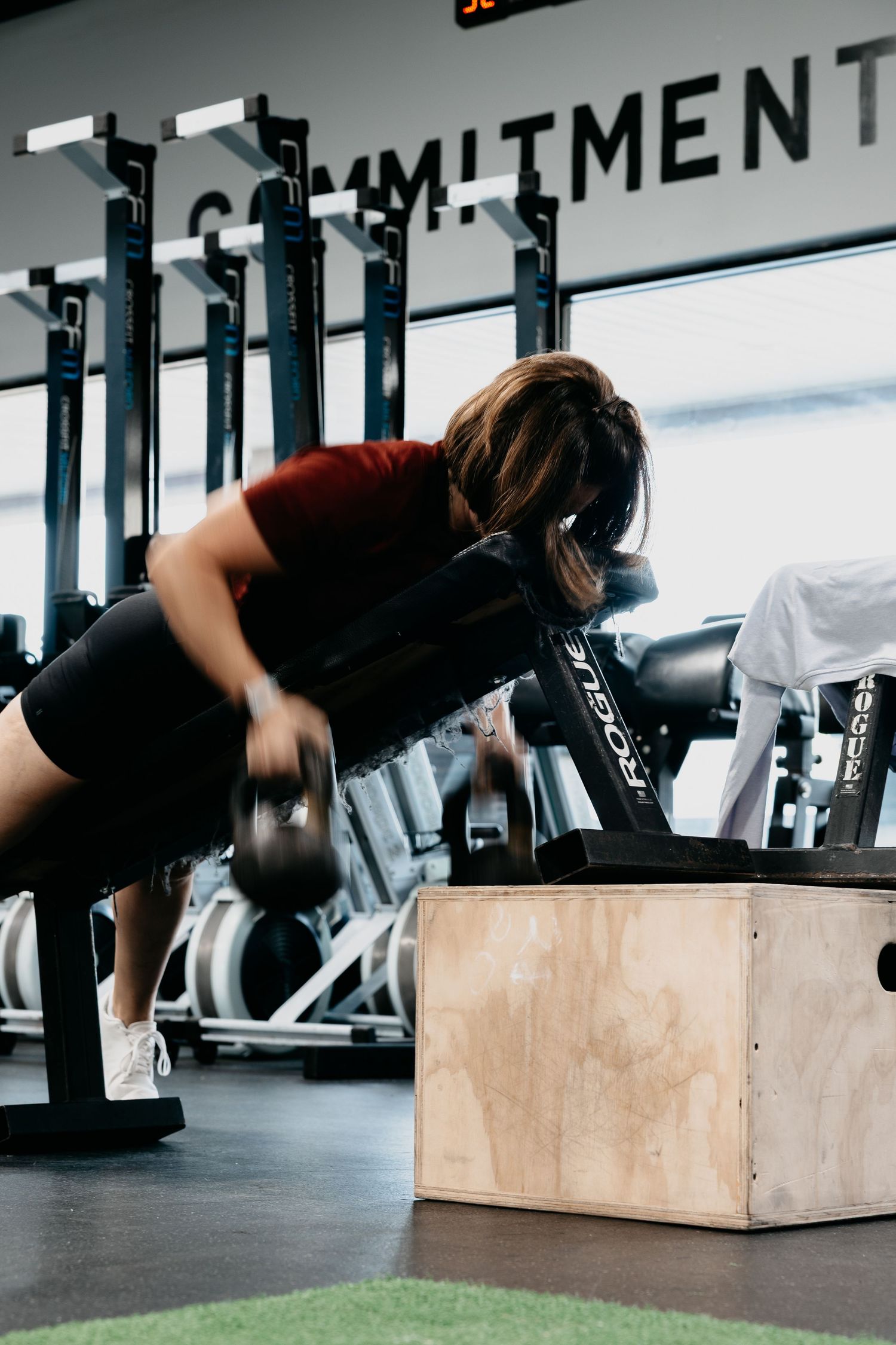 Woman performing dumbbell rows on a wooden plyo box in a modern gym with motivational signage.