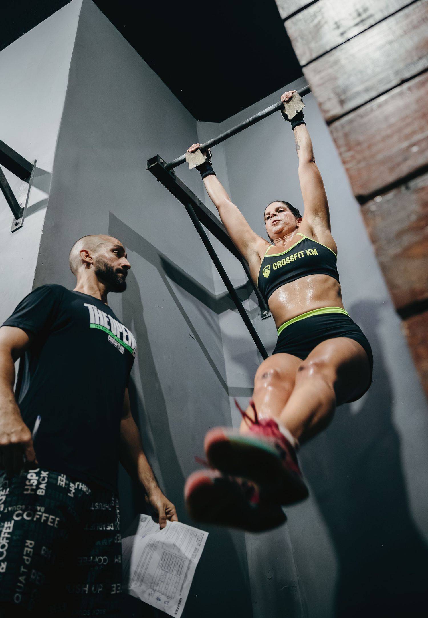 Woman performs pull-ups on bar at CrossFit gym while male coach observes during intense training session.