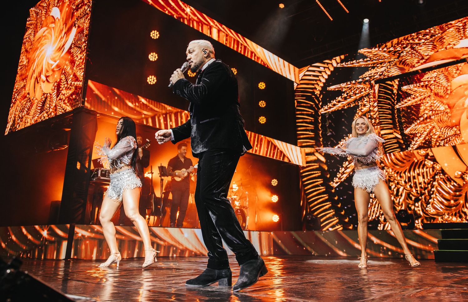 Male performer in black suit sings on stage with dancers in silver outfits and dramatic orange lighting backdrop.