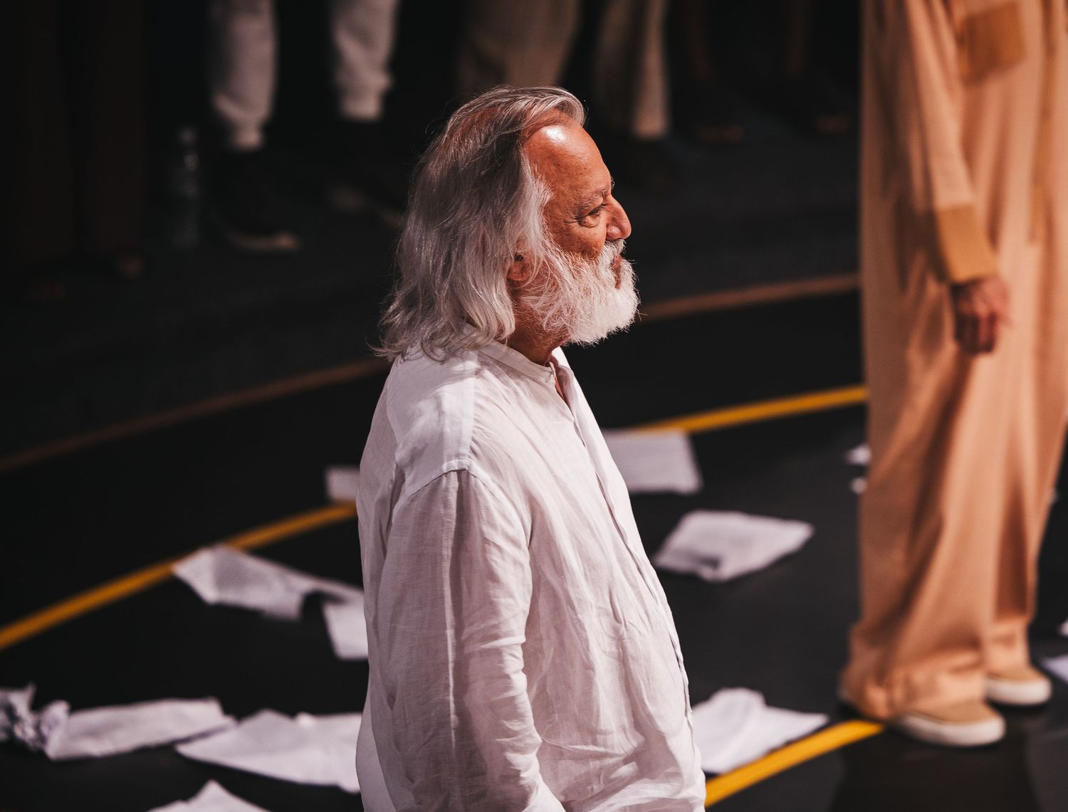 Elderly man with long white hair and beard in white linen shirt stands among scattered papers on dark stage floor.