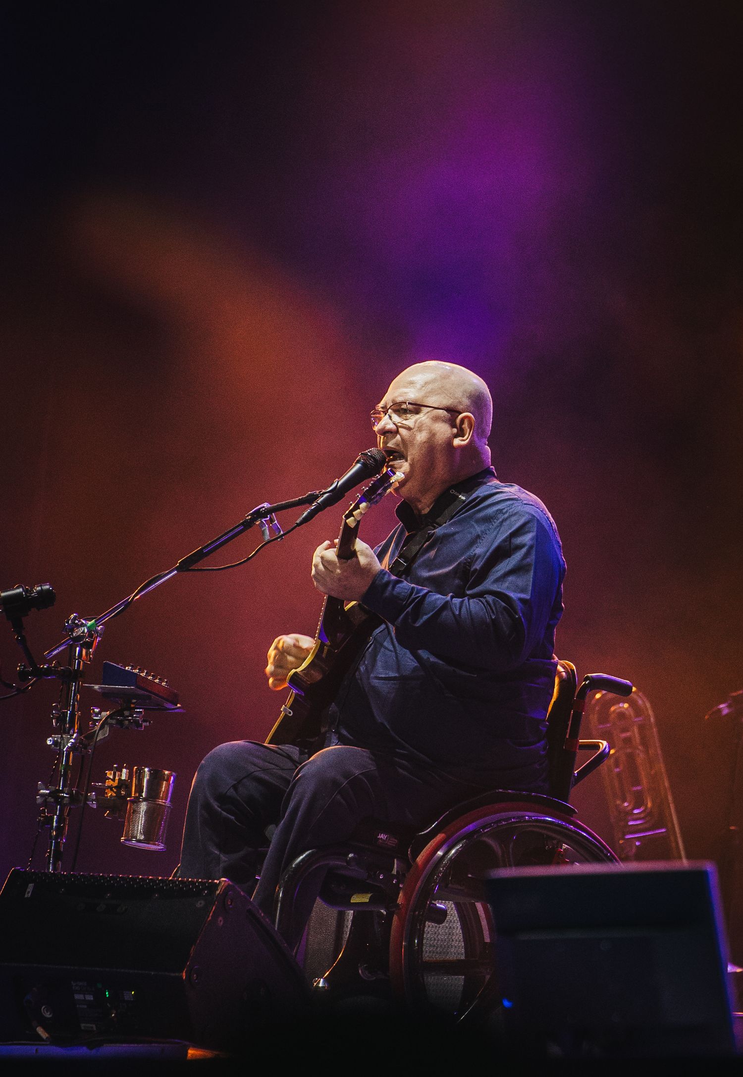 Bald musician in wheelchair plays guitar and sings into microphone on stage with colorful purple and red lighting.