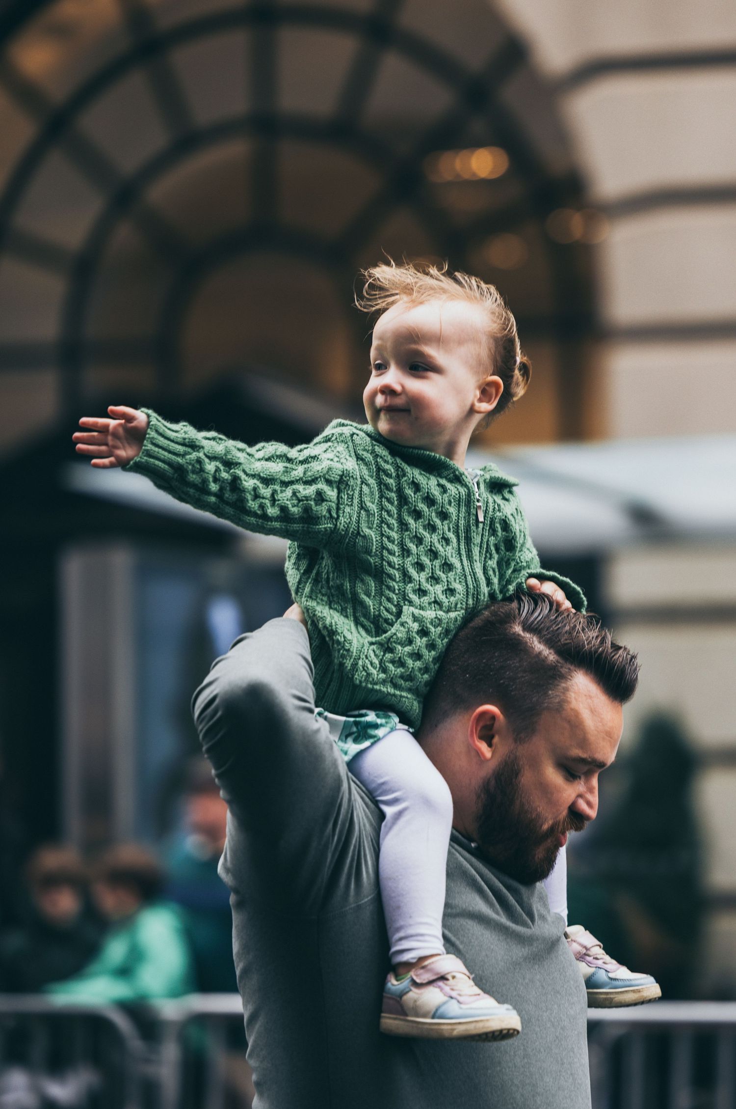 Smiling toddler in green knit sweater sitting on a mans shoulders with arms outstretched outdoors.