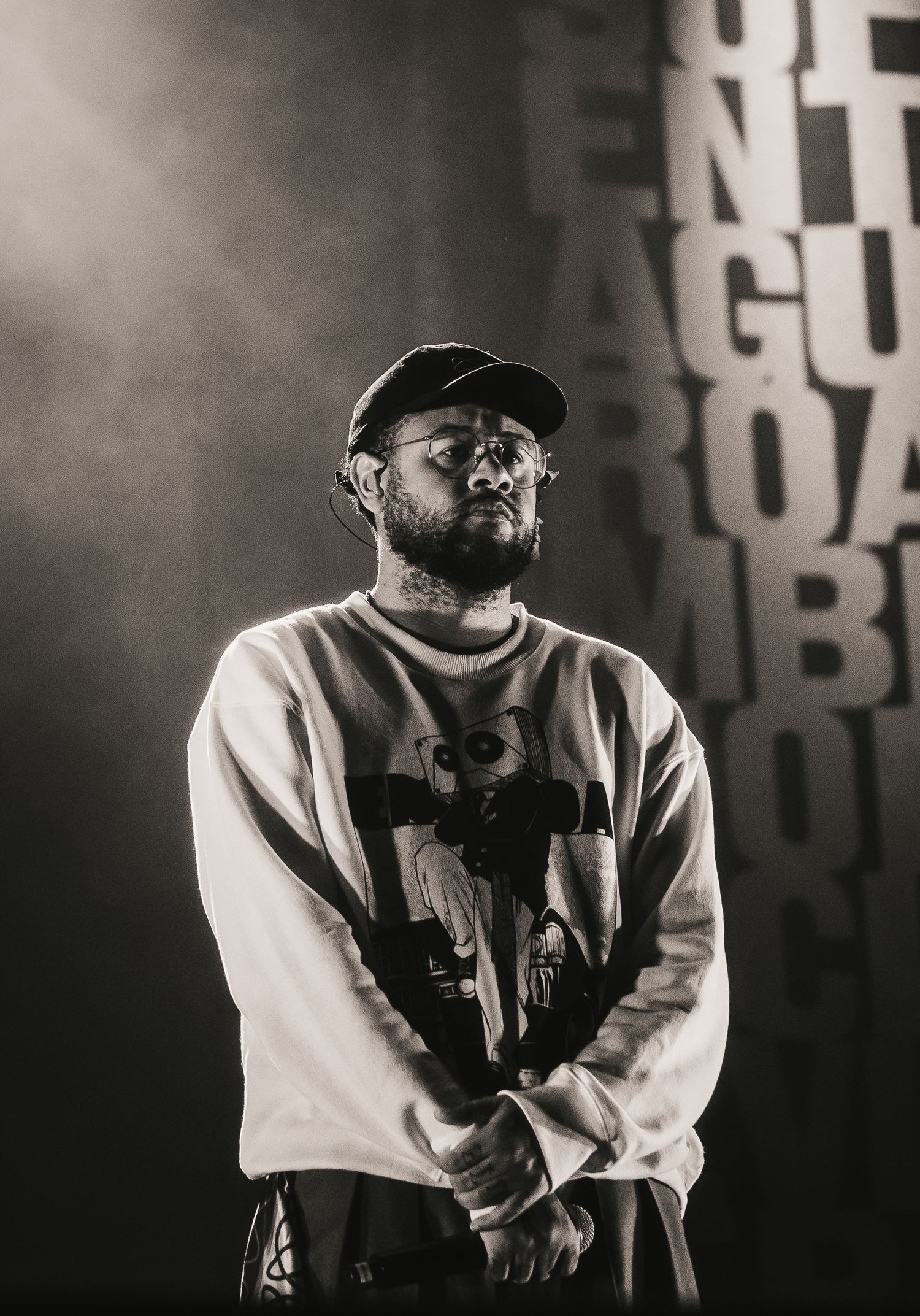 Black and white portrait of bearded male rapper on stage wearing cap and graphic sweatshirt.