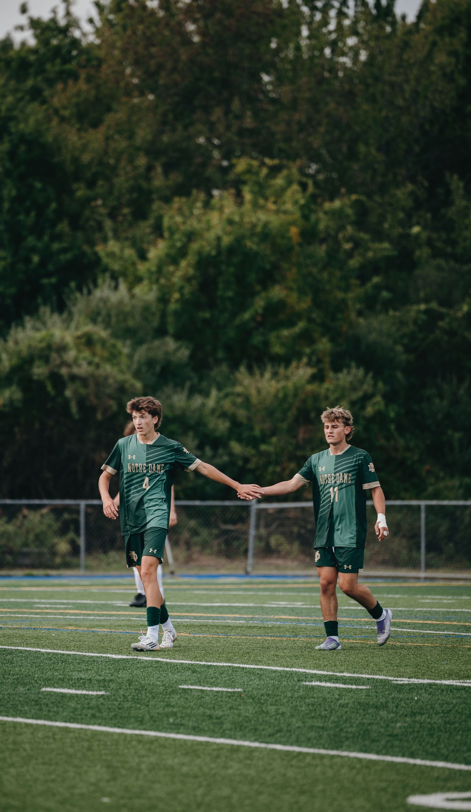 Two youth soccer players in green jerseys high-five on a turf field during a match.
