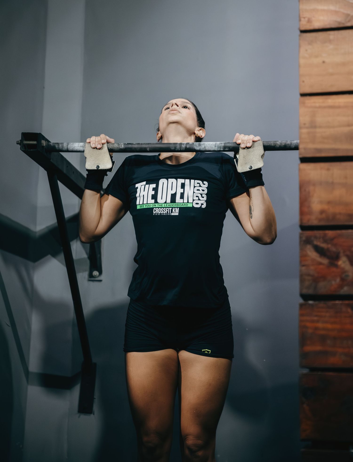Woman performing pull-ups in a gym wearing a black CrossFit Open t-shirt and workout gloves.