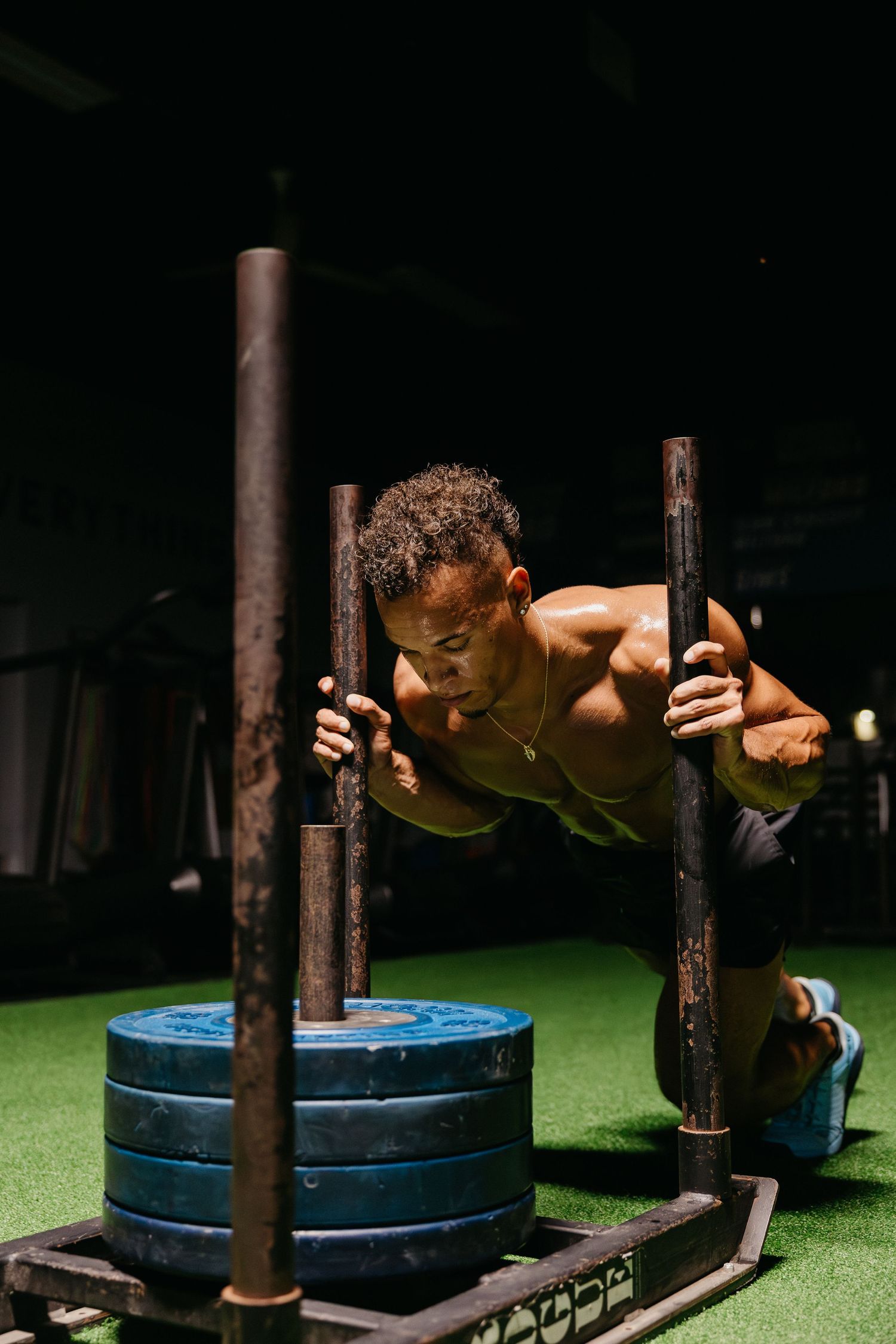 Muscular shirtless athlete pushing a weighted sled in a dark gym during an intense CrossFit training session.