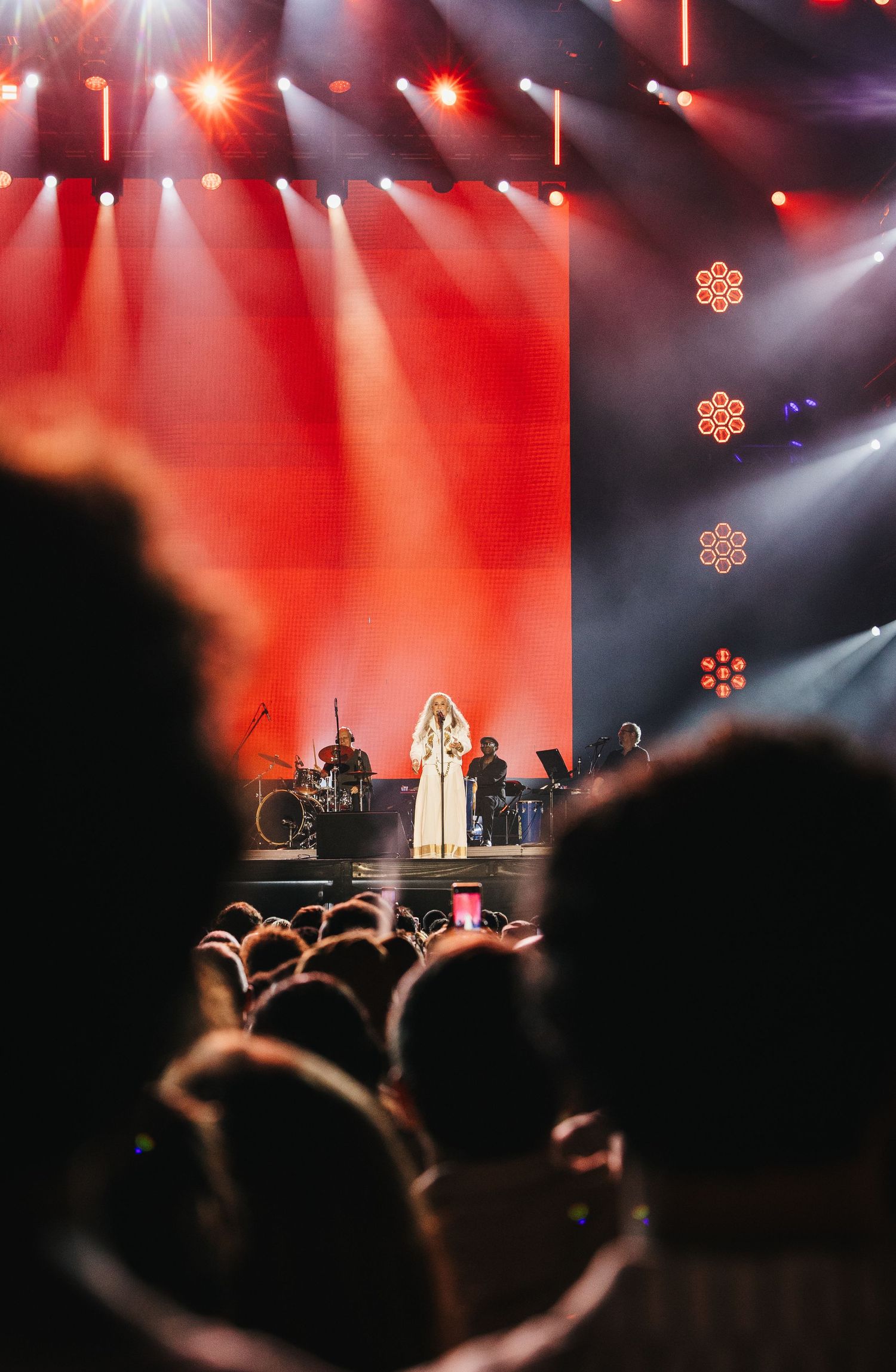 Female singer performs on a red-lit stage before a large crowd at a live concert event.
