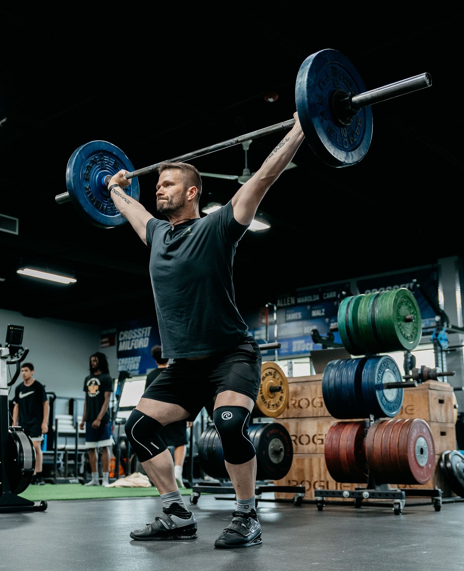 Athlete performing overhead squat with barbell in a CrossFit gym, wearing knee sleeves and black workout attire.