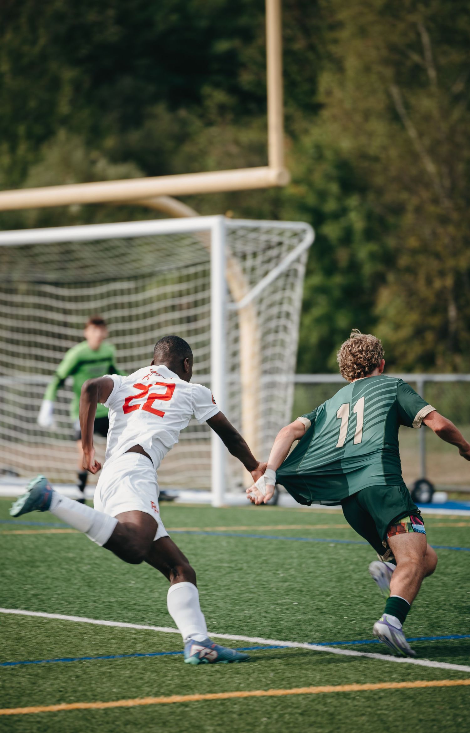 Soccer players #22 and #11 compete on turf field with goalkeeper defending goal during intense match.