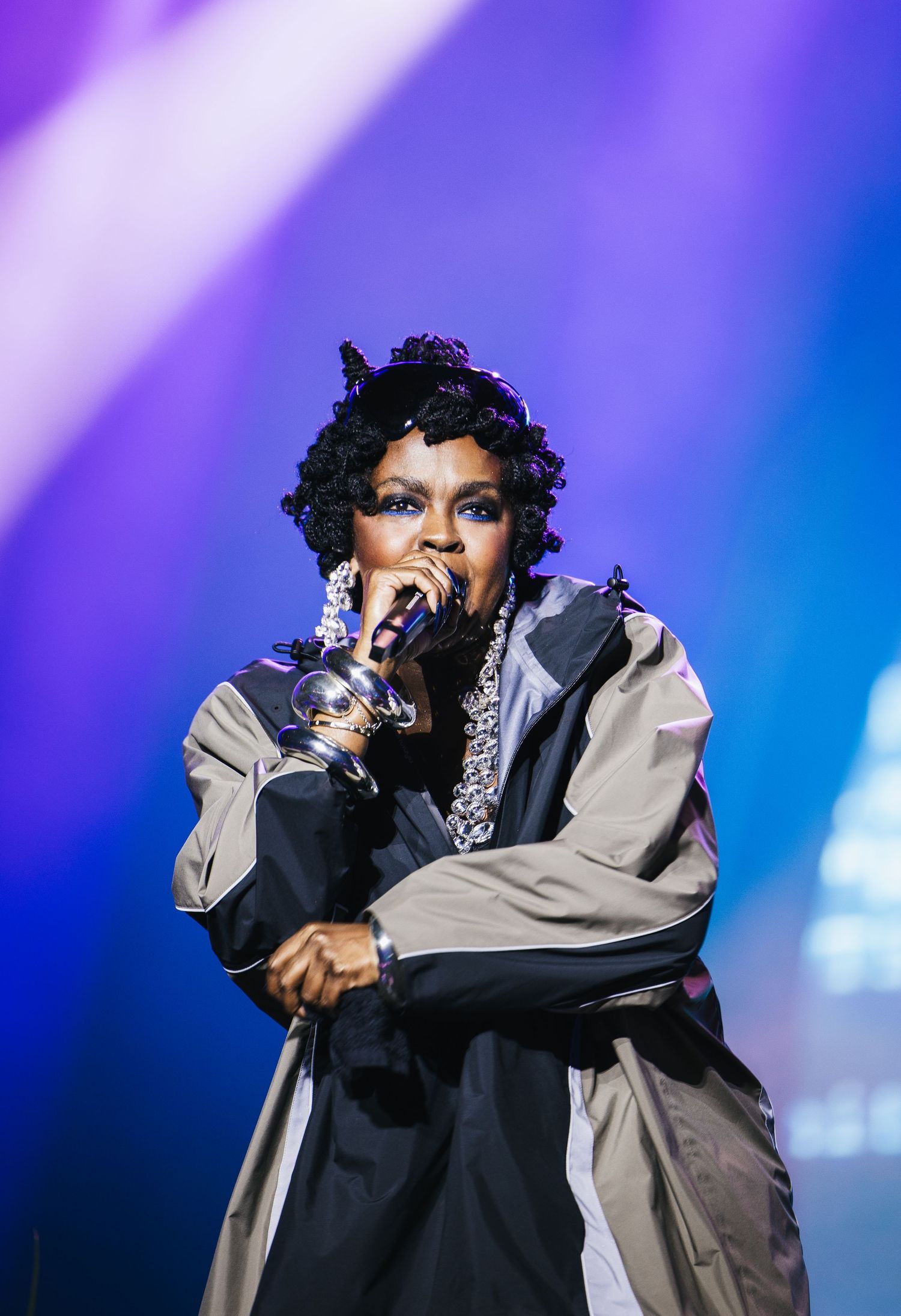 Female performer singing on stage with microphone, wearing gray jacket and chain necklace under purple stage lighting.