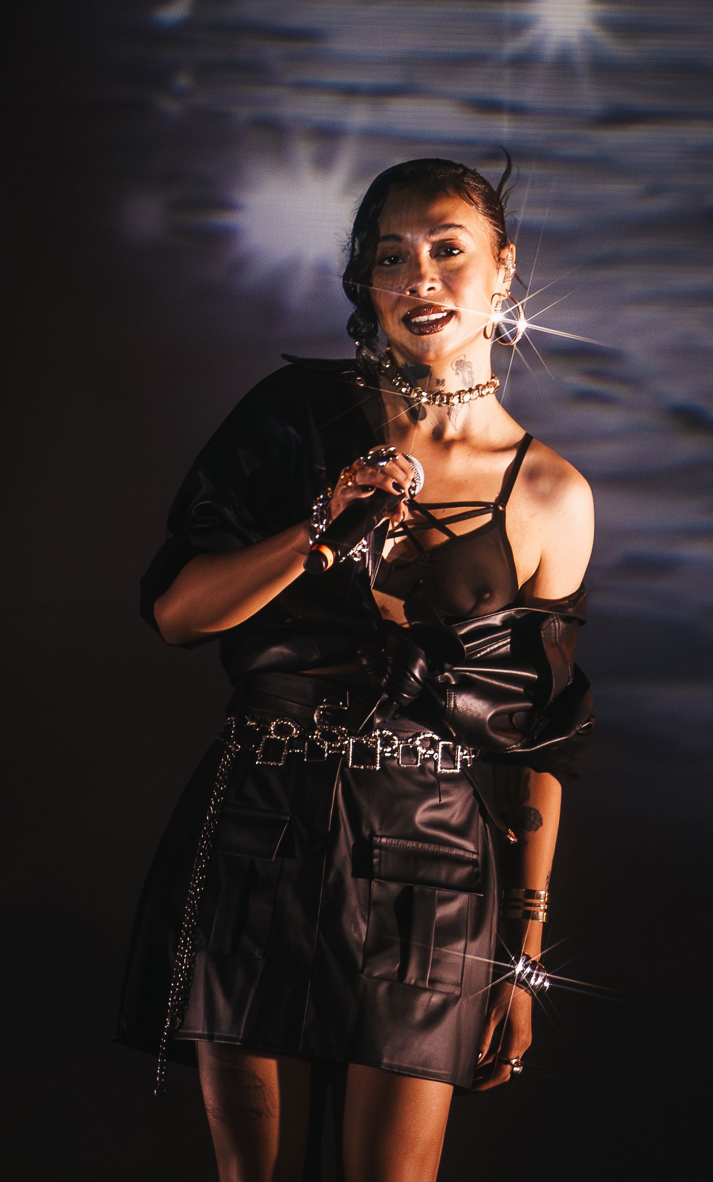 Female performer in black leather outfit with silver jewelry sings on stage under bright spotlight.