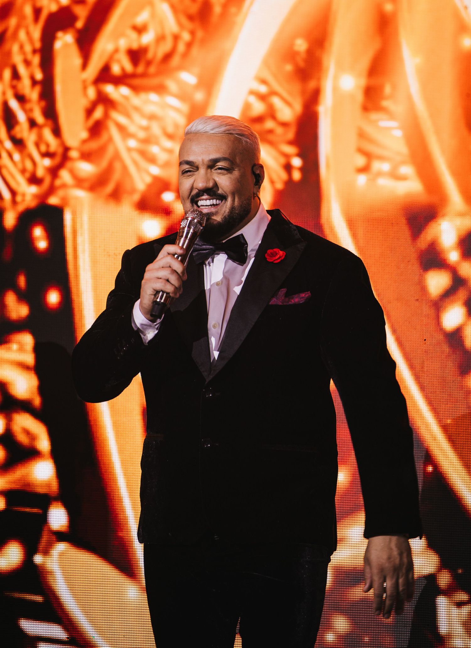 Man in black tuxedo with red boutonniere smiles while holding microphone on stage with orange backdrop.