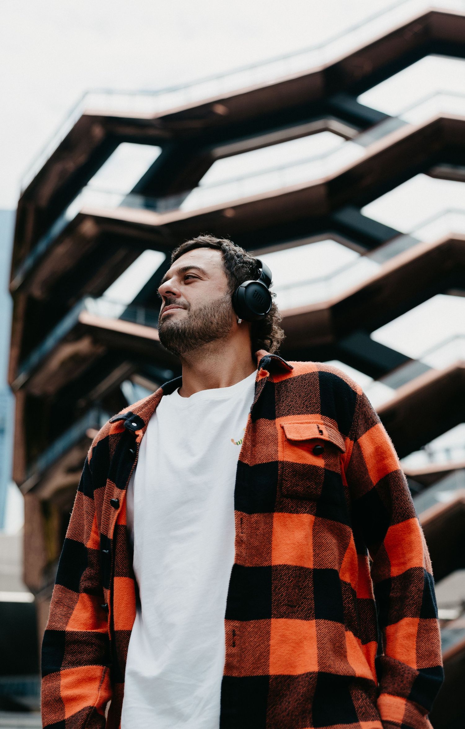 Young man with headphones wearing plaid flannel shirt standing outdoors near modern architecture.