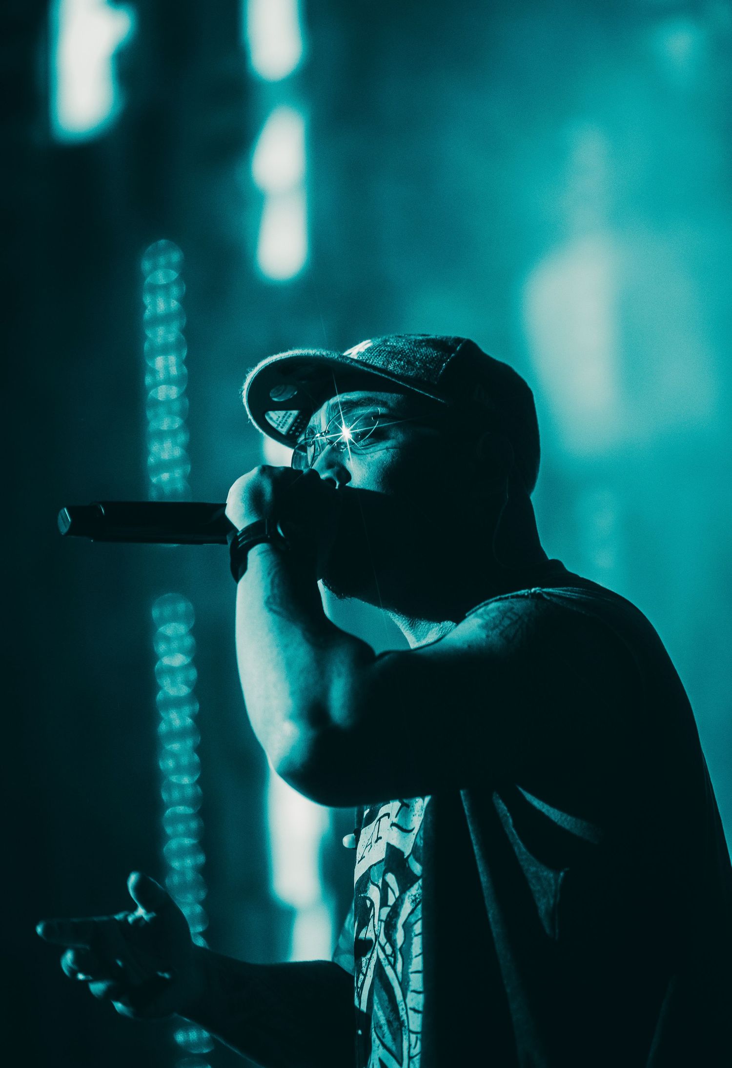 Rapper performing on stage under teal blue lighting, holding microphone close while wearing a cap.