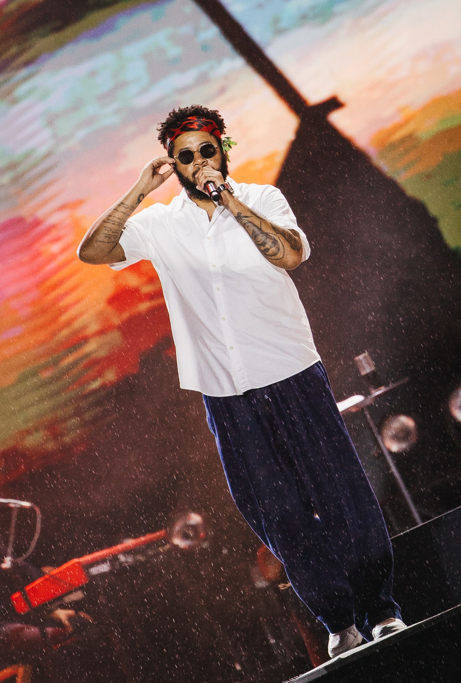 Tattooed male performer in white shirt and sunglasses singing on stage with colorful backdrop.