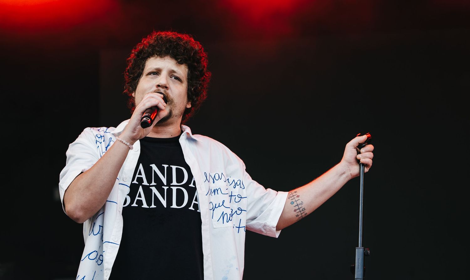 Singer performing on stage wearing a white jacket over a black band t-shirt, holding a microphone.