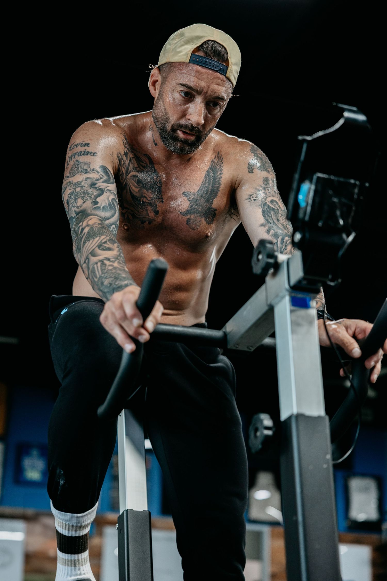 Tattooed shirtless man with prosthetic leg working out on exercise bike in gym wearing cap.