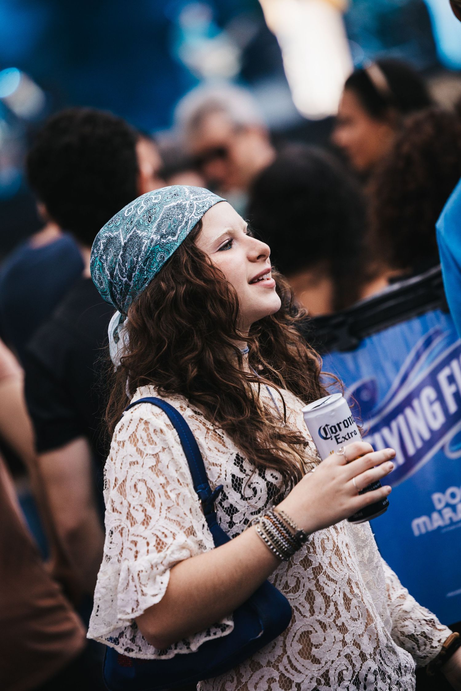Young woman in lace top and bandana holding Corona beer at outdoor music festival.