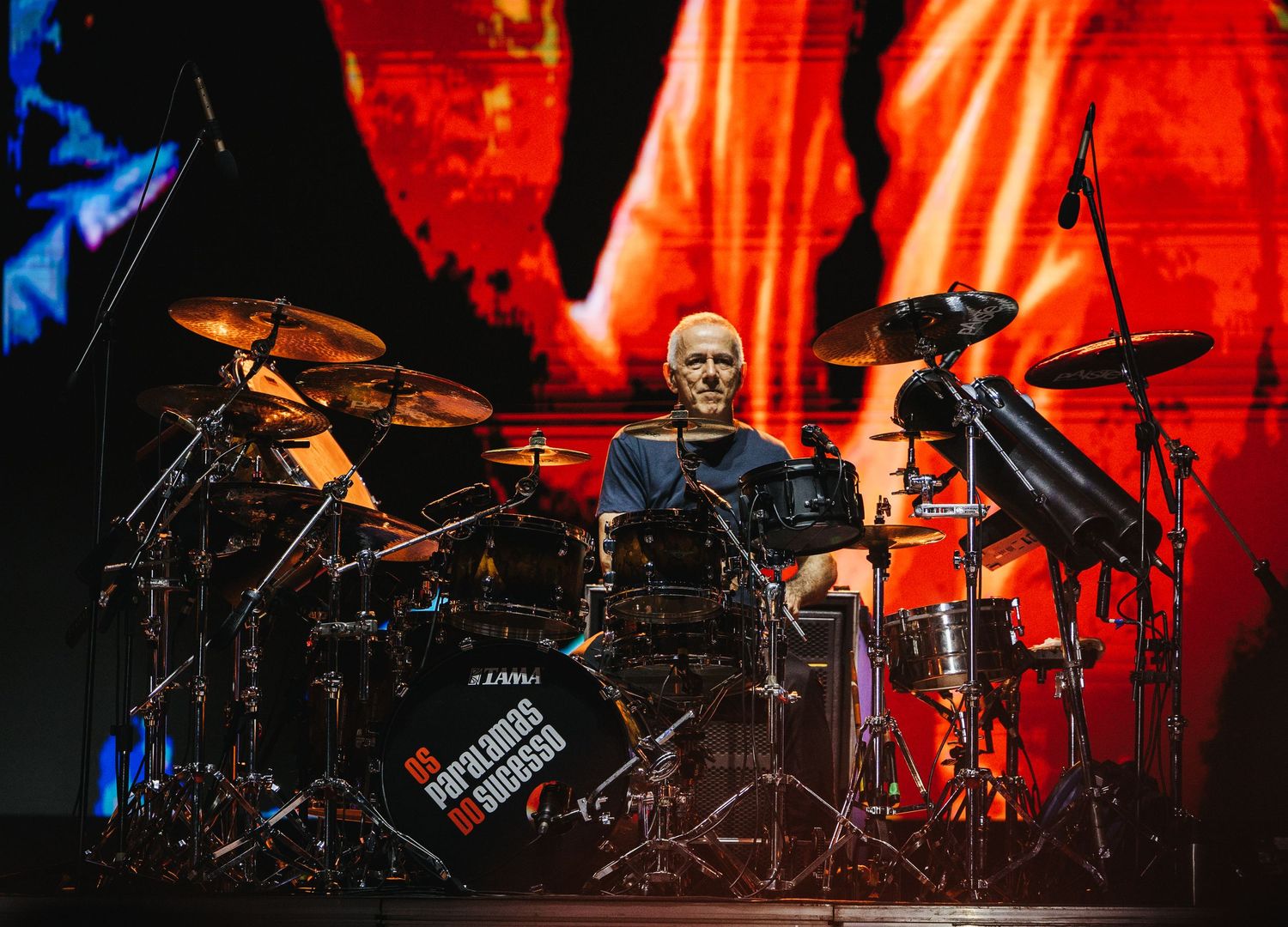 Drummer performing on stage at a live concert with cymbals and drum kit, vibrant red backdrop lighting.
