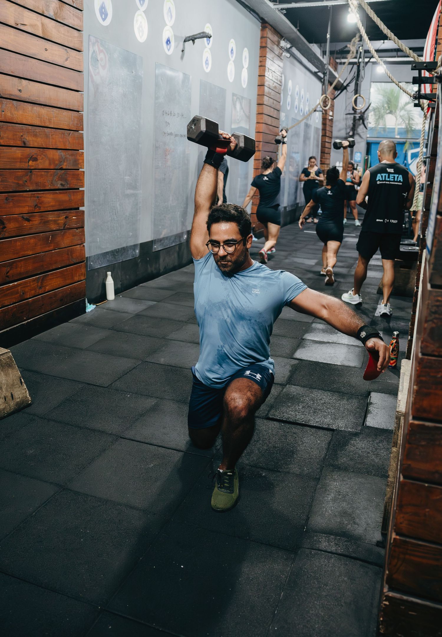 Man performing a dumbbell overhead lunge in a CrossFit gym with other athletes training in the background.