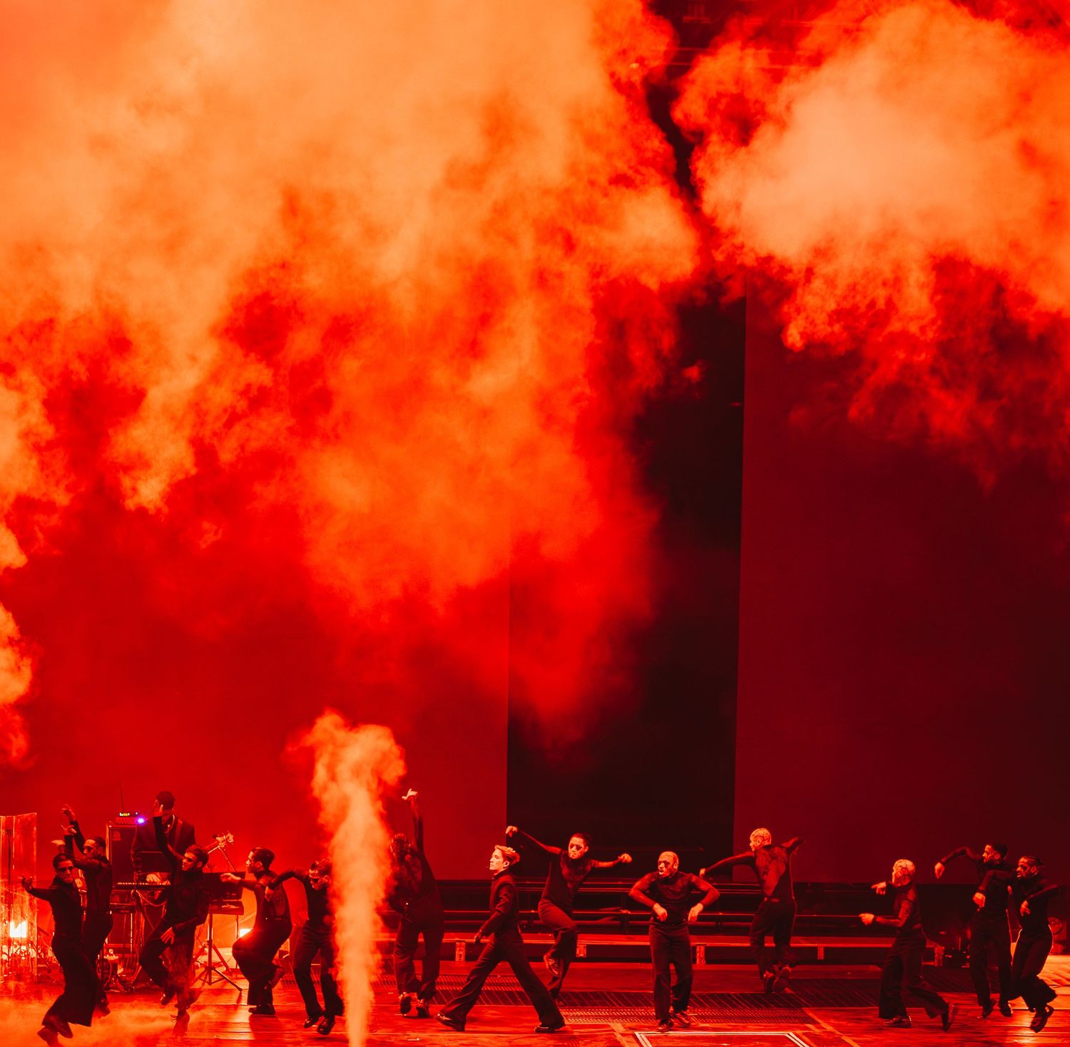 Dramatic red smoke fills a concert stage as performers move through intense pyrotechnic effects.