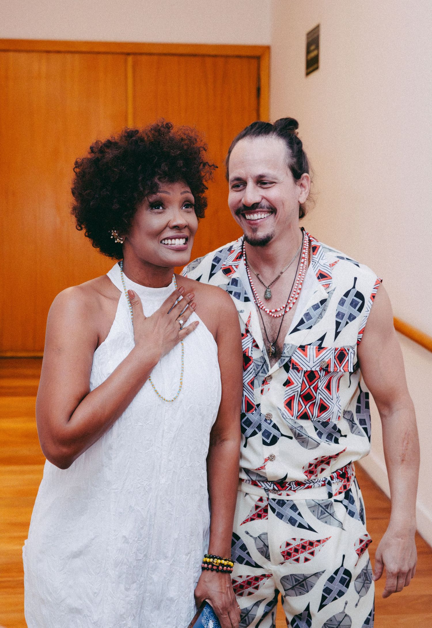 A smiling woman with an afro and a man in a patterned outfit pose together in a bright indoor space.