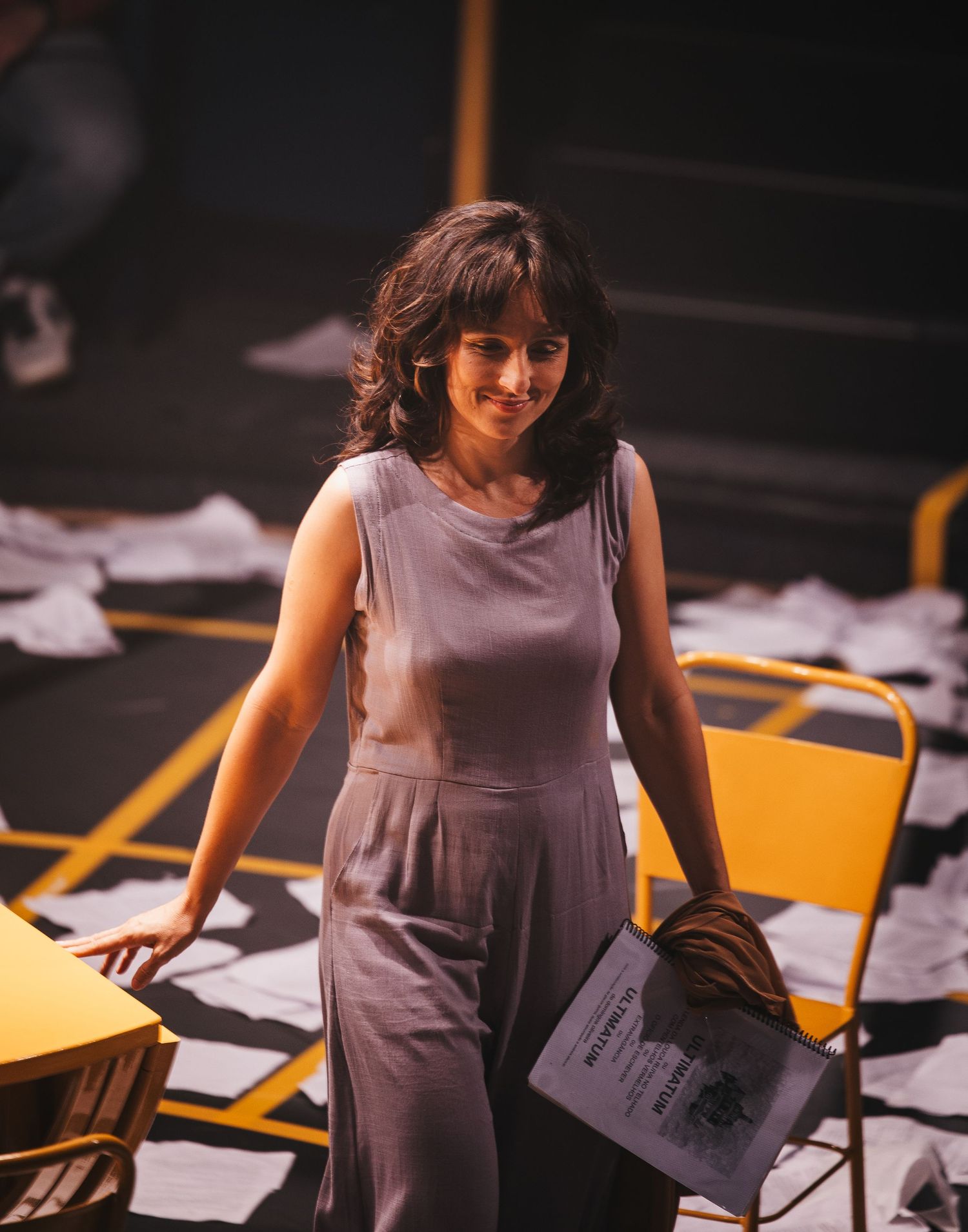 Woman in gray dress holding newspaper stands among scattered papers and yellow chairs on a stage.