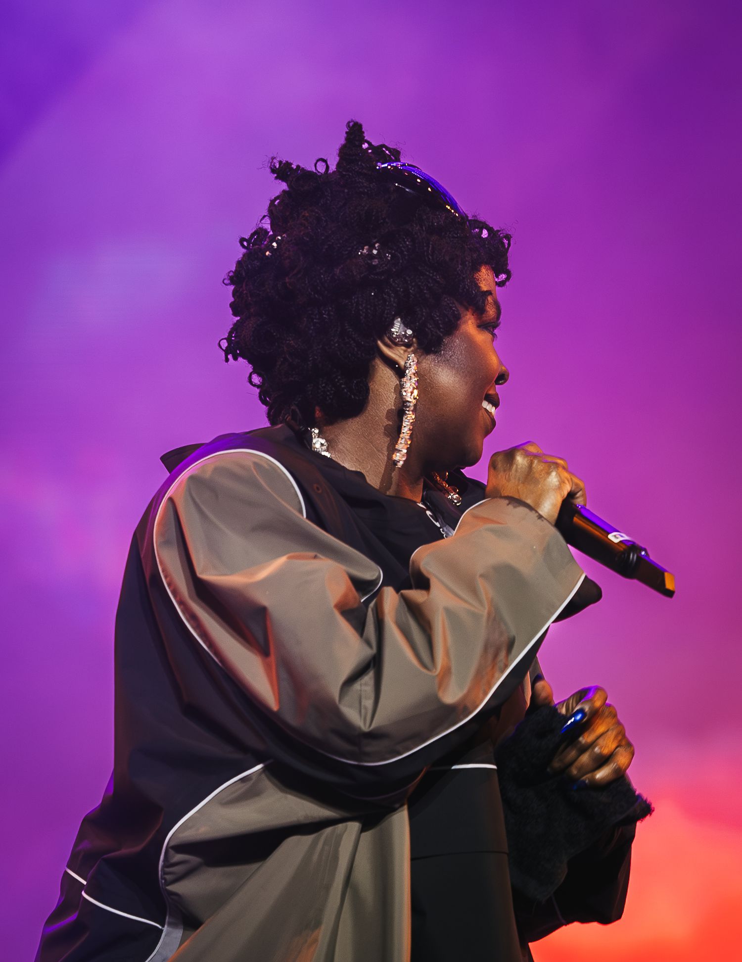 A performer sings into a microphone on stage under purple and pink lights wearing a gray and black jacket.