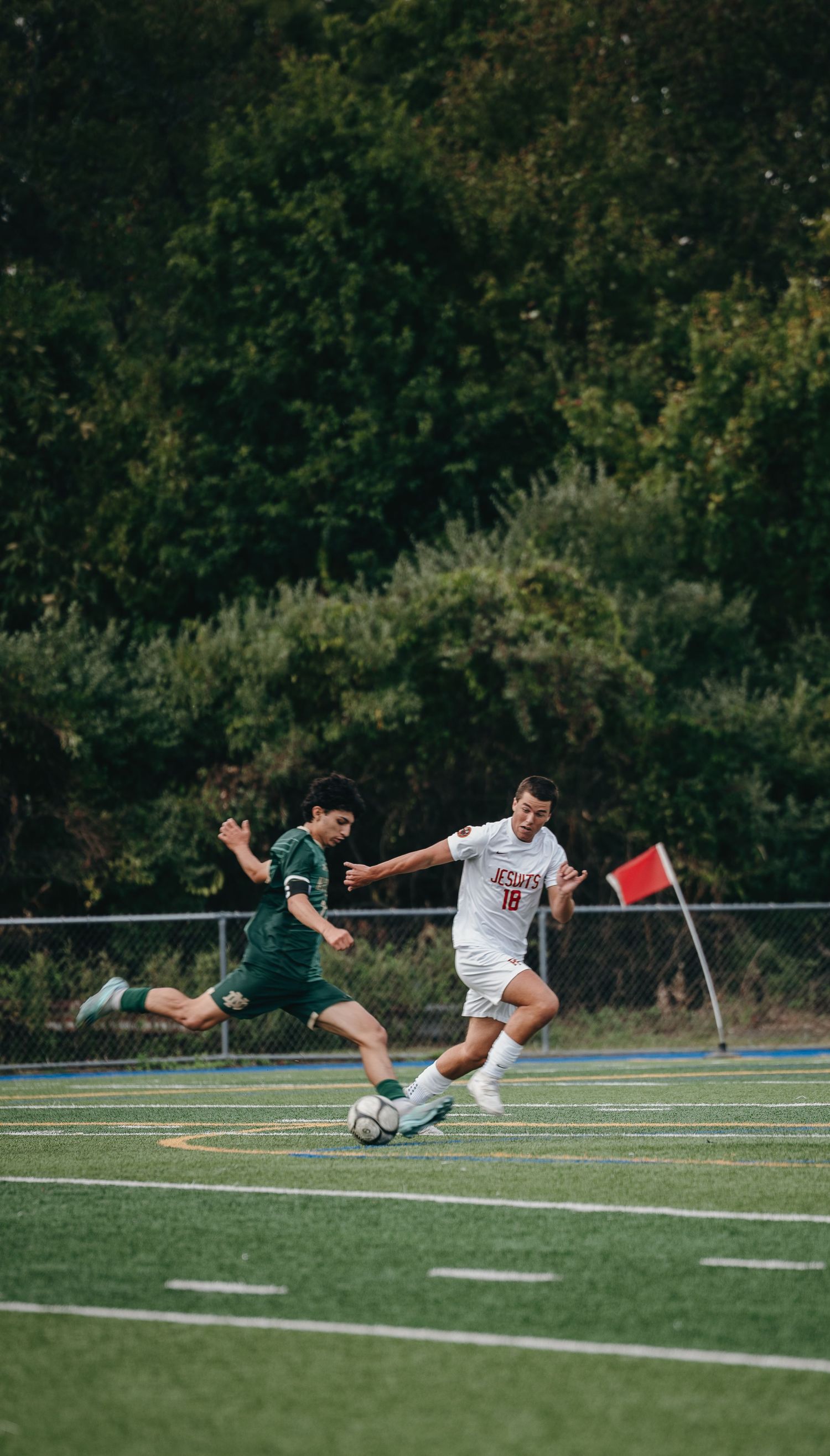 Two soccer players compete for the ball on a green turf field during a match, with trees in the background.