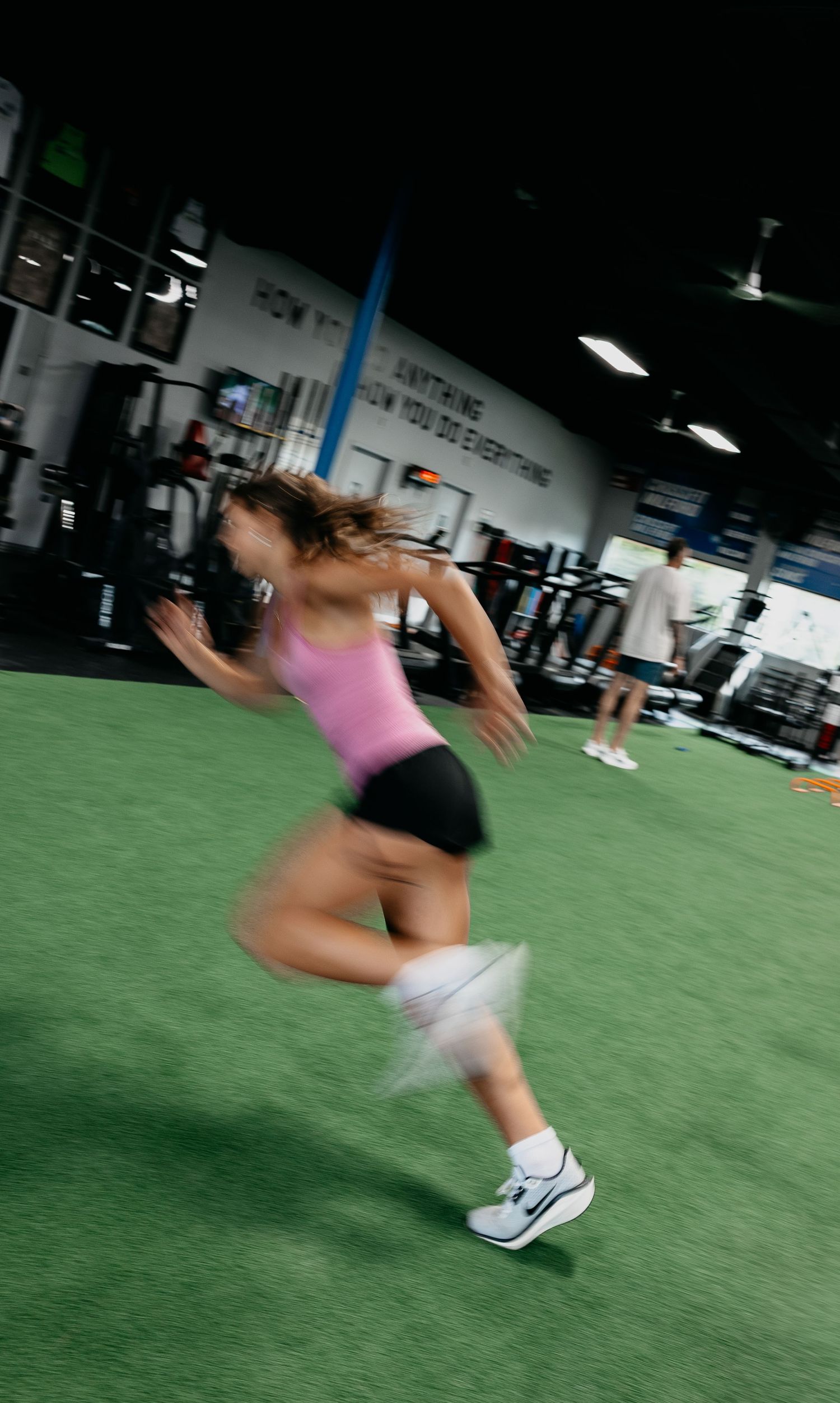Woman sprinting across green turf in a gym wearing pink top and black shorts, motion blur capturing her athletic speed.
